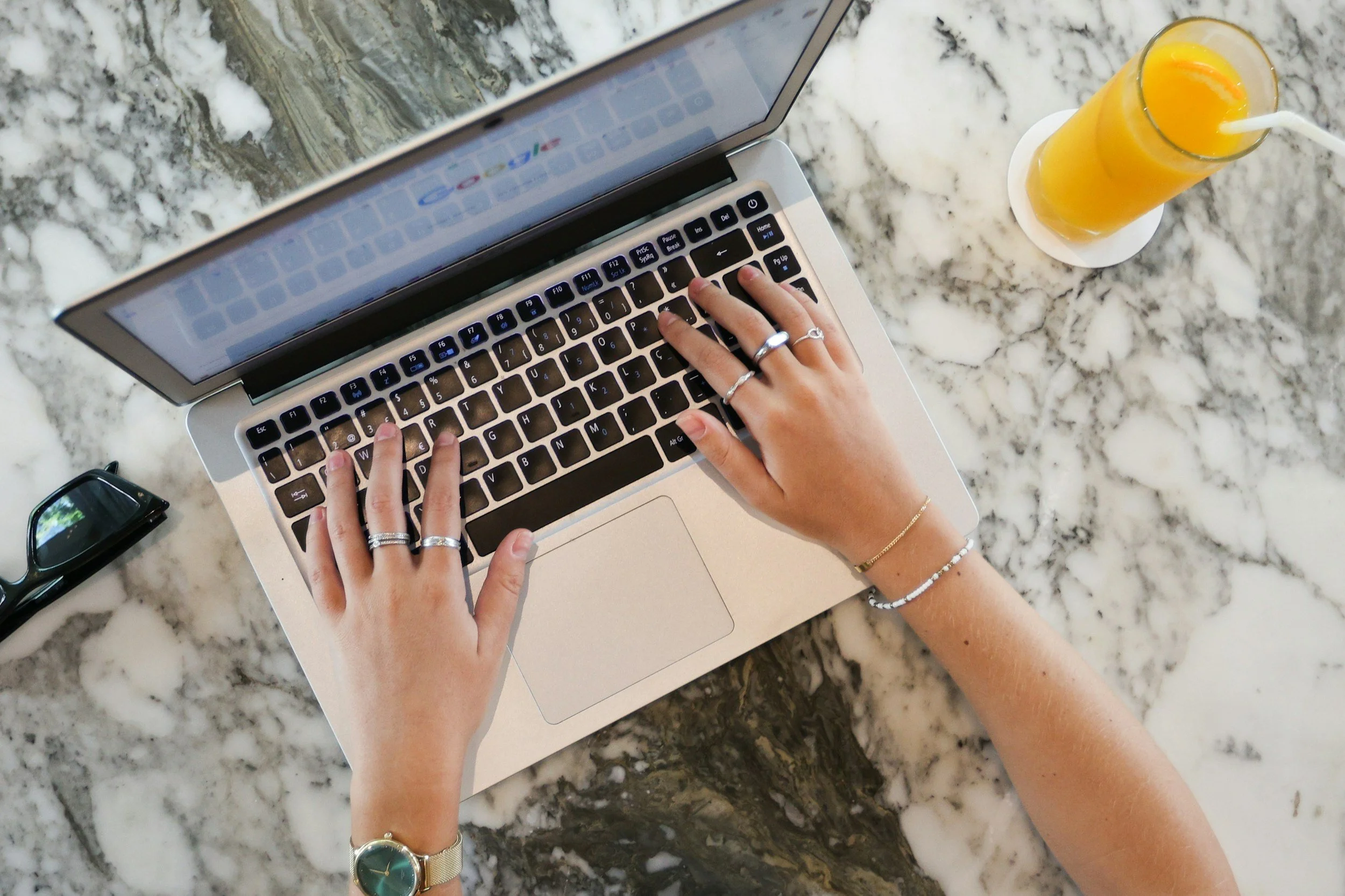 Person typing on a laptop keyboard at a marble table with a glass of orange juice, sunglasses, and jewelry on their hands and wrist.