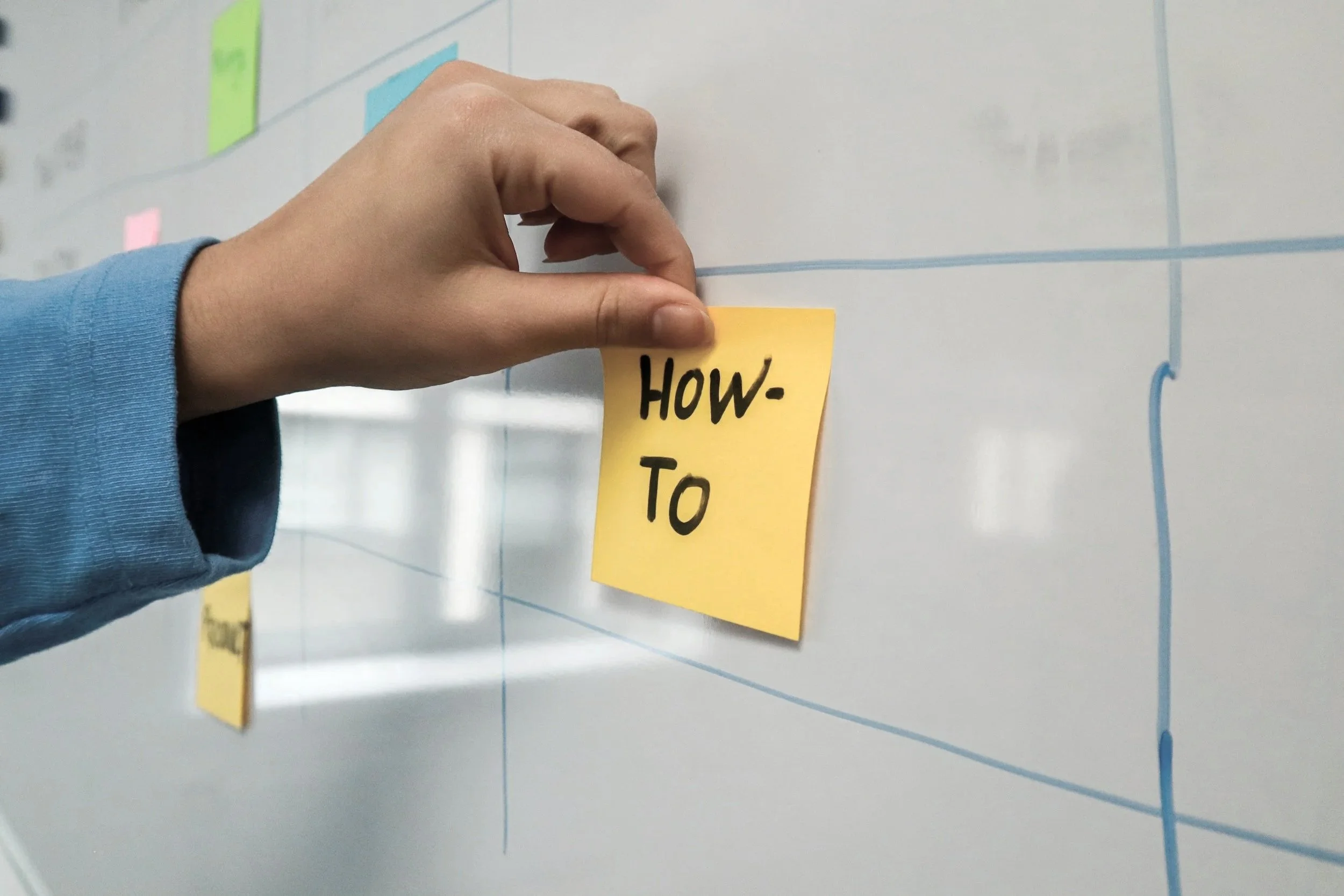 Hand placing a yellow sticky note labeled 'HOW-TO' on a whiteboard with blue grid lines.