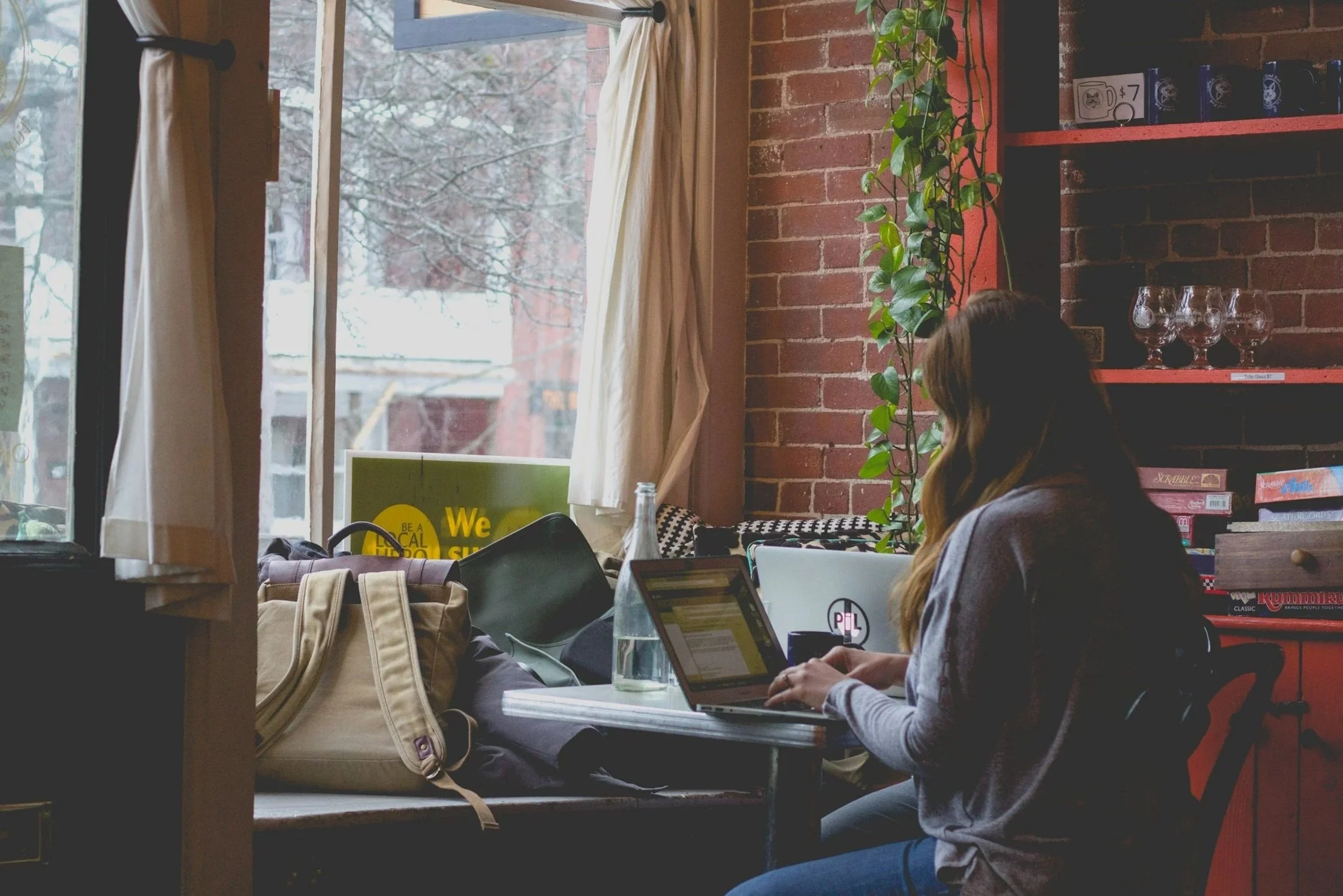 A woman working on a laptop at a small table in a cozy coffee shop or cafe, with a large window, brick wall, and shelves behind her.