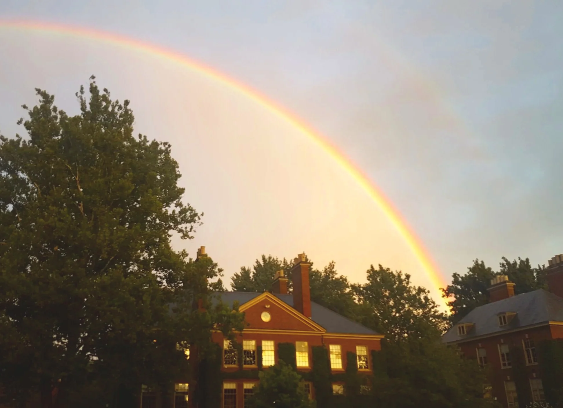 A rainbow arching over a brick school building with tall trees in the foreground during sunset.
