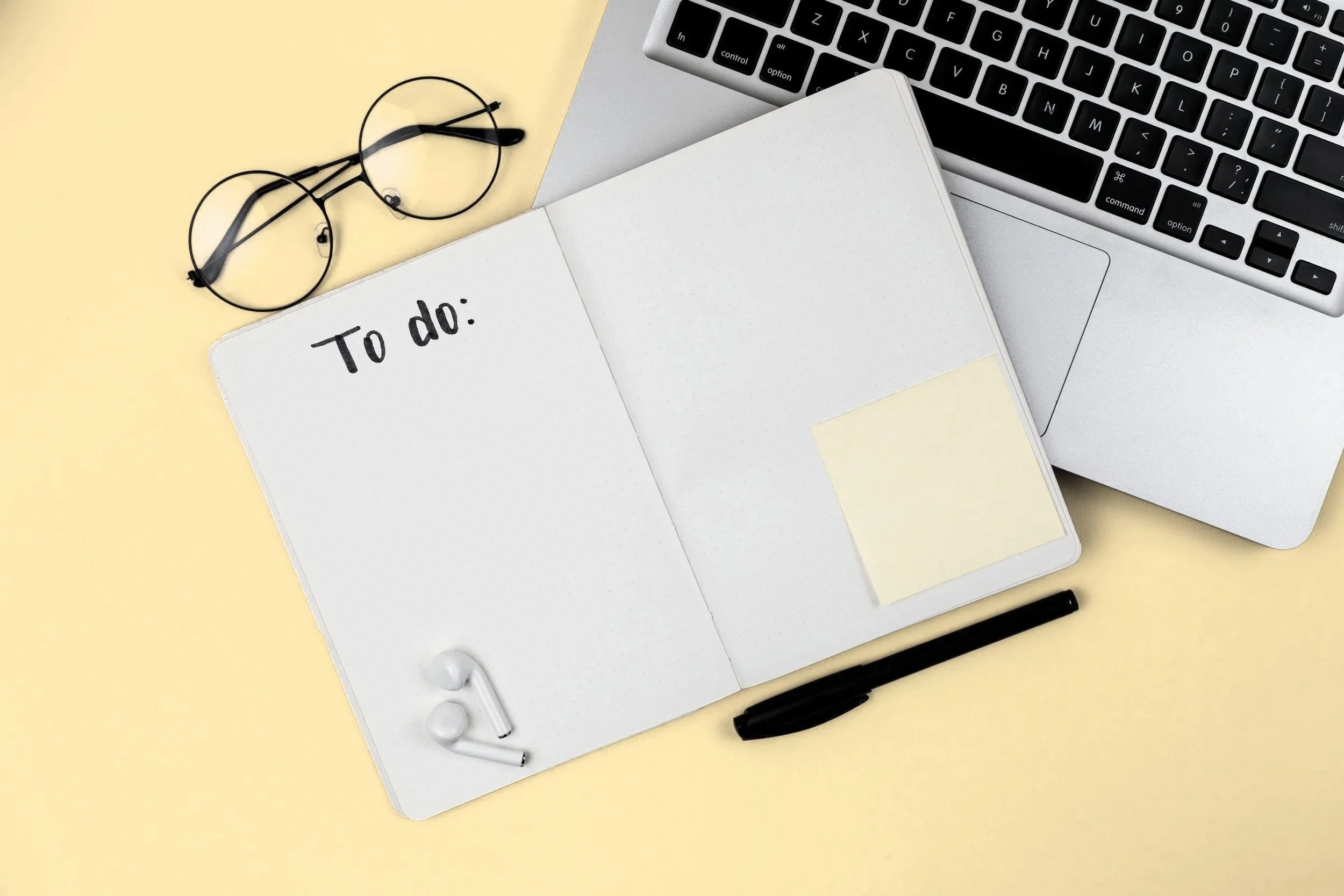 Flat lay of a workspace on a yellow background including a silver laptop with black keyboard, an open dotted notebook with a "To do:" list written on the left page, a yellow sticky note on the right page, black pen, black and white earbuds, a pair of black-rimmed glasses, and a black marker.