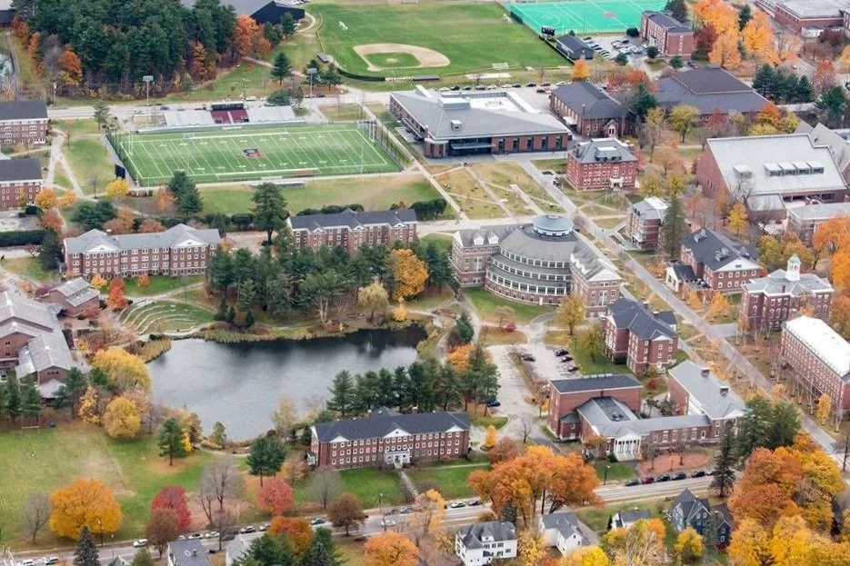 An aerial view of a college campus during fall with colorful trees, a pond, a football field, and various academic buildings and dormitories.