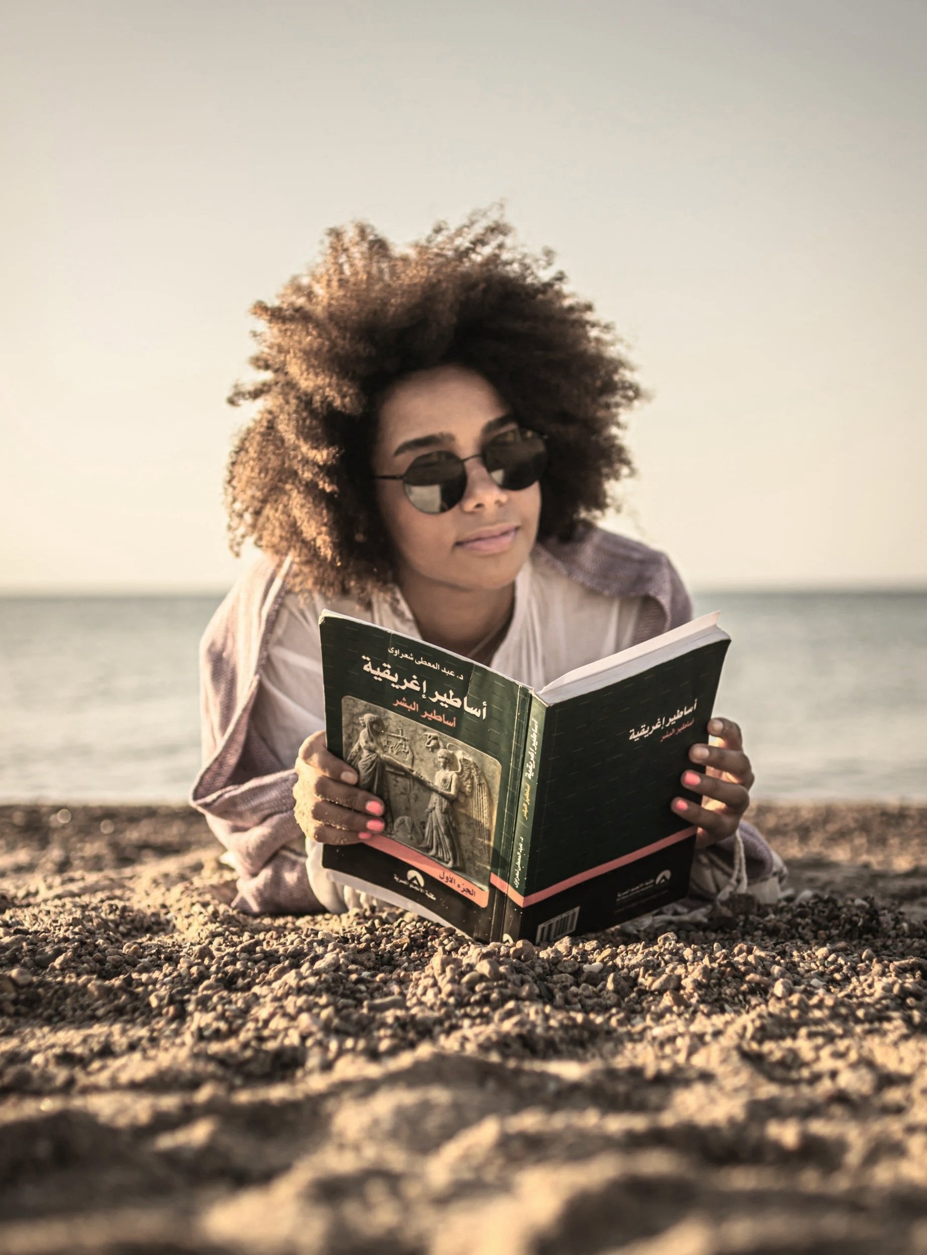 A woman with curly hair and sunglasses reading a book on a beach.