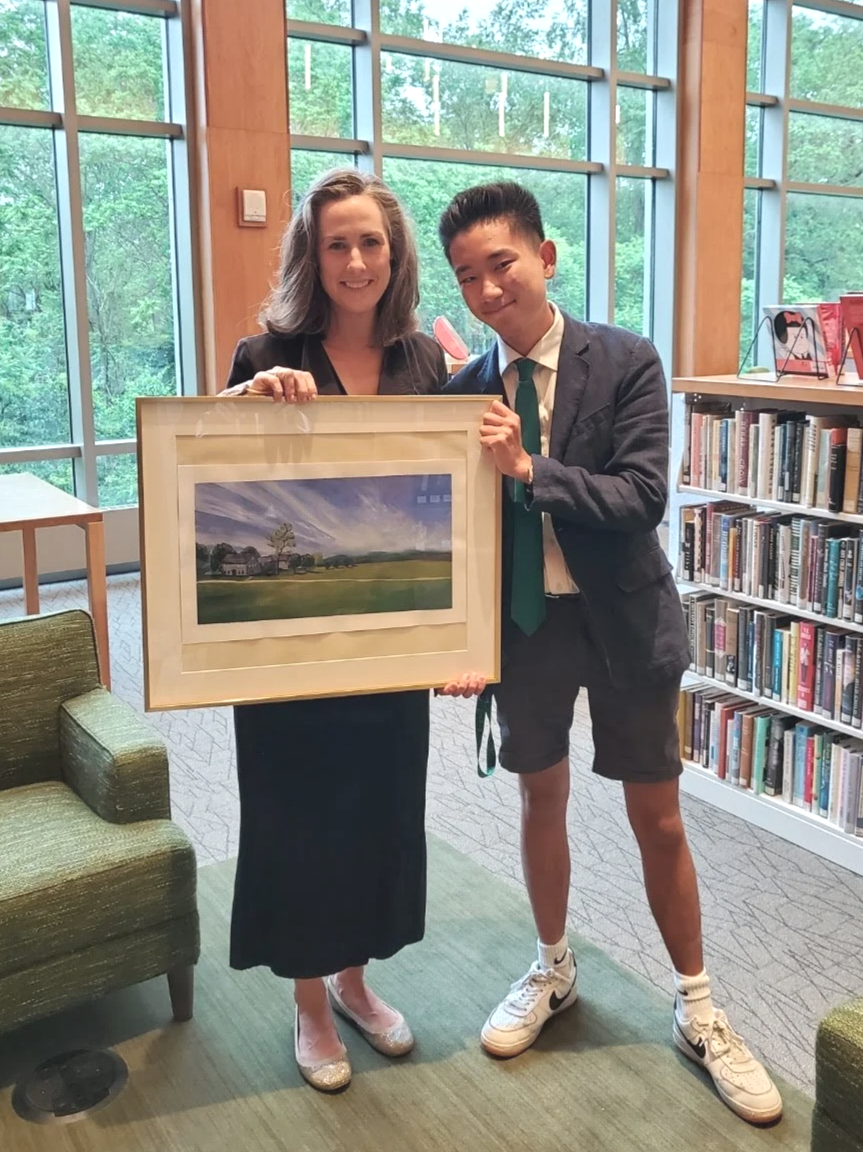A woman and a teenage boy holding a framed landscape painting inside a library. The woman wears a black dress and heels, the boy wears a blazer, white shirt, tie, shorts, and sneakers. Large windows with greenery outside are in the background.