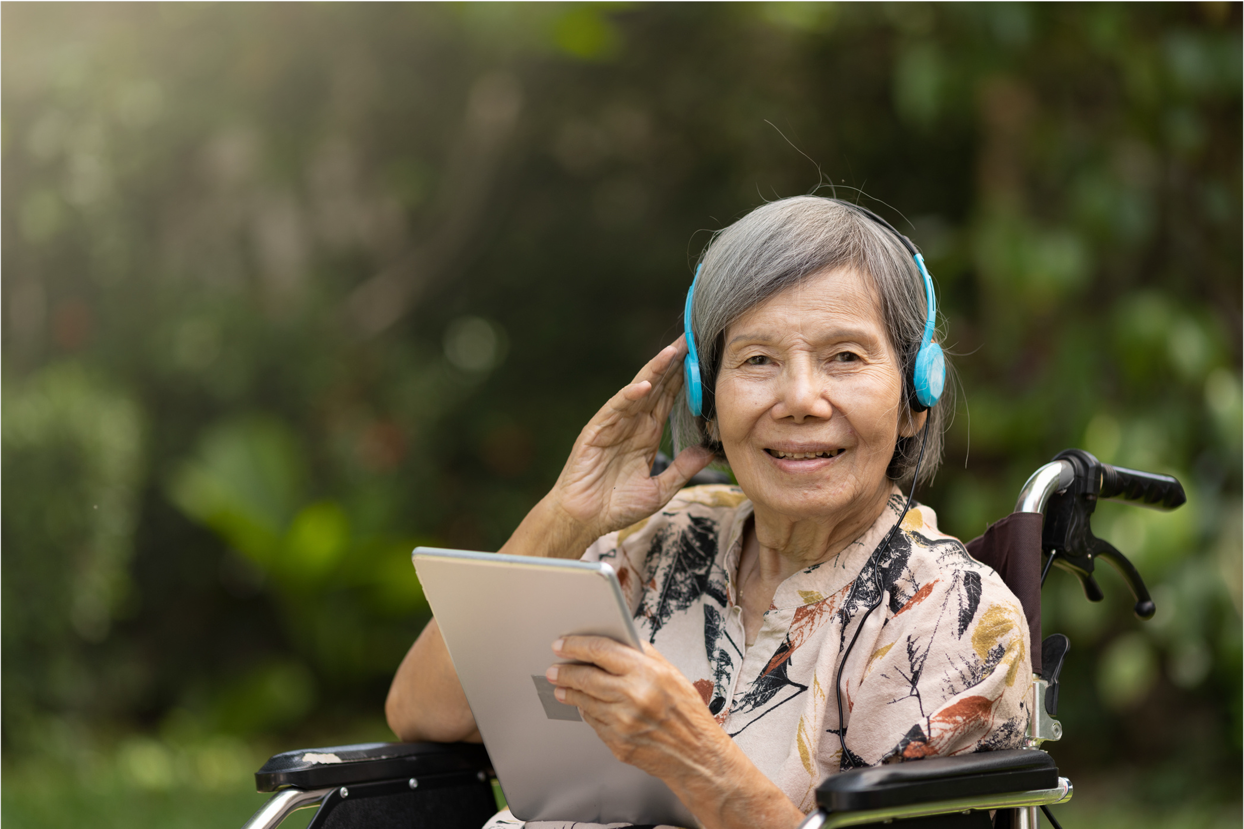 An elderly woman with gray hair sitting in a wheelchair outdoors, wearing blue headphones, holding a tablet, smiling, and enjoying music or an audio.