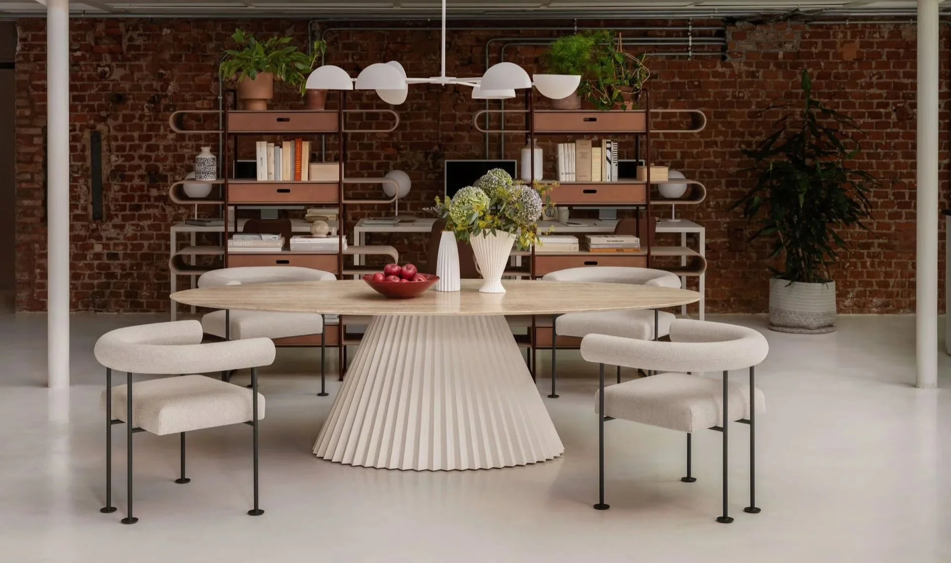 Modern dining room with a round wooden table, six beige upholstered chairs, a white fluted base, a white vase with flowers, a red bowl of apples, and a brick wall with shelving units holding books and plants.