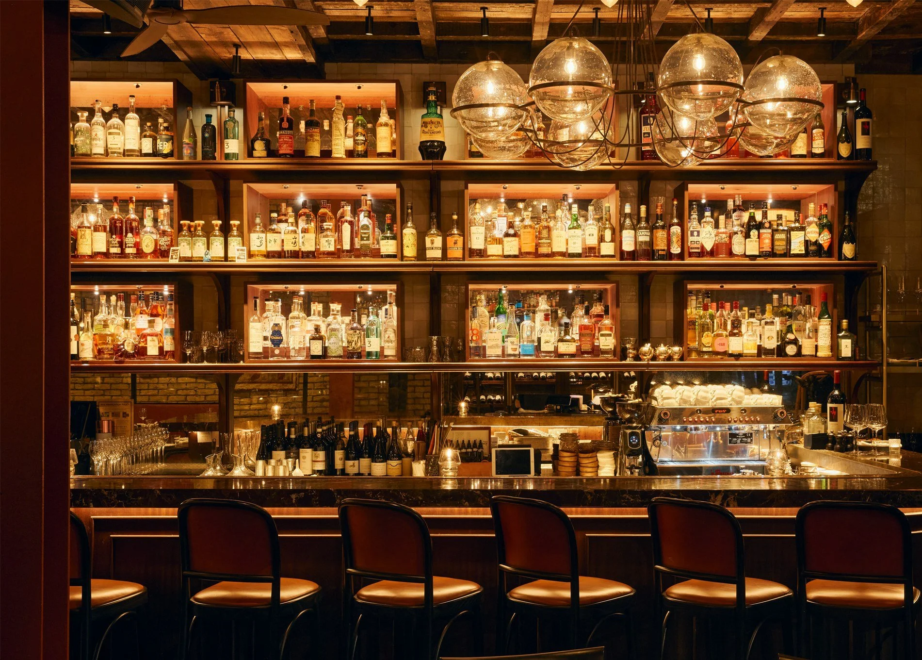 Bar with shelves filled with various liquor bottles, illuminated with warm lighting, and dark chairs lined up in front.
