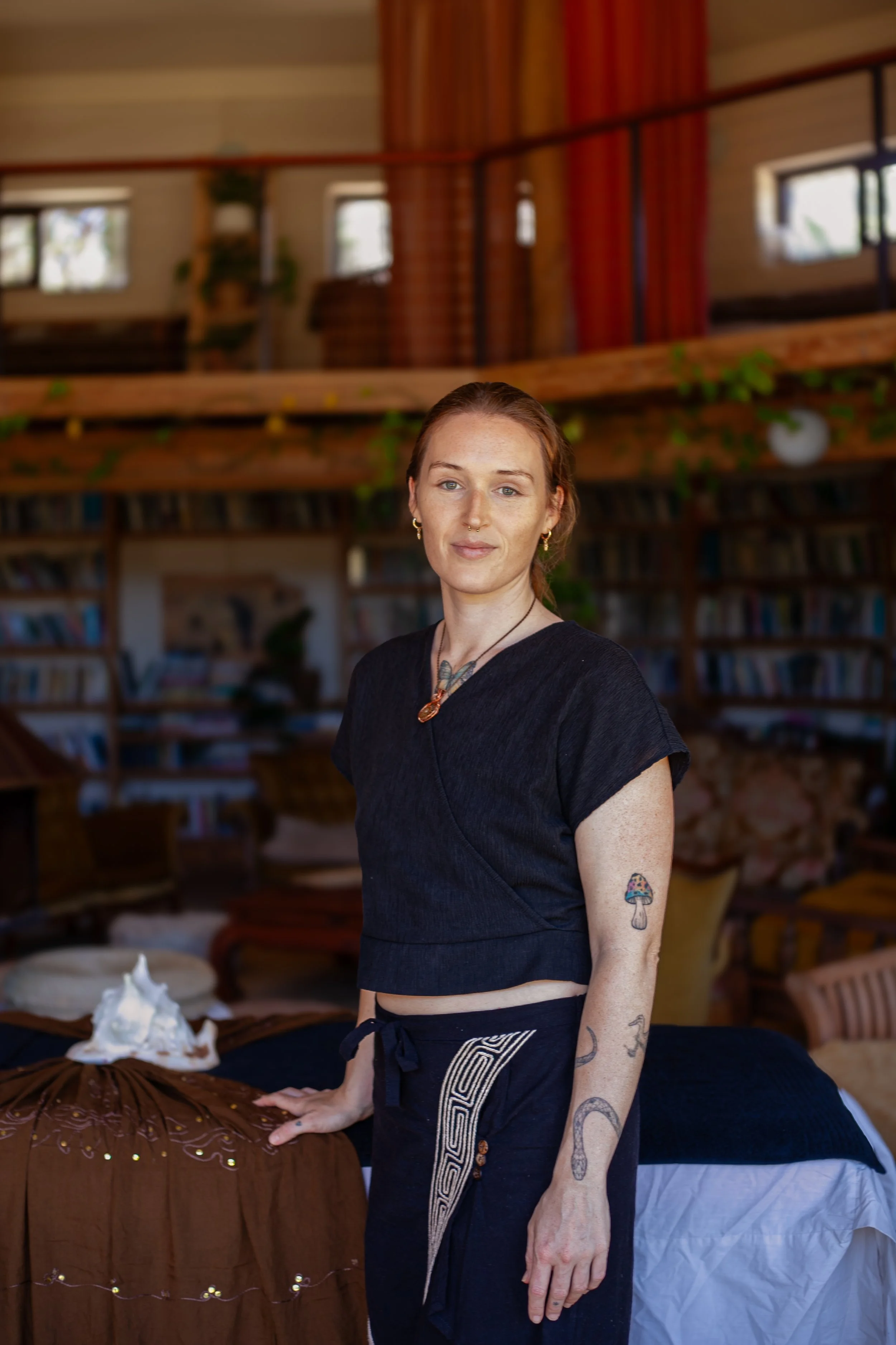 A woman with reddish hair and tattoos on her arms standing in a cozy, wood-paneled room with bookshelves, plants, and a bed with a blanket in the background.