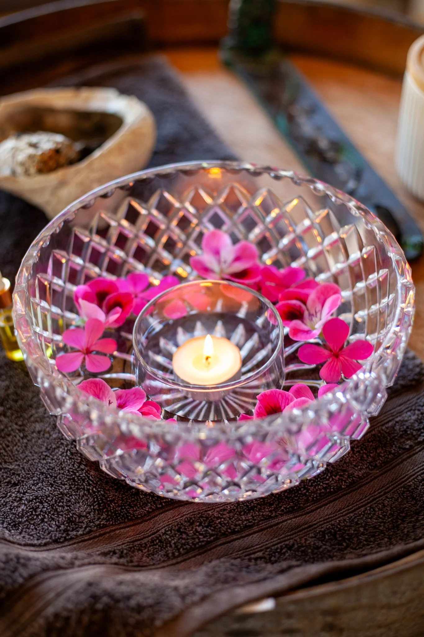 A decorative glass bowl filled with pink flower petals, with a small lit tealight candle at the center, on a dark cloth surface.