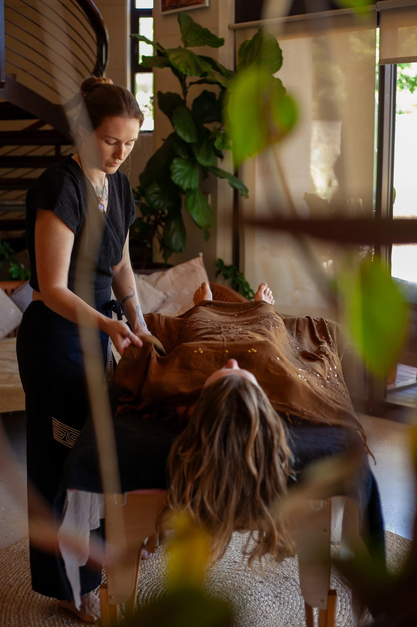 A woman giving a massage to a person lying on a massage table inside a cozy room with large windows and lush plants.