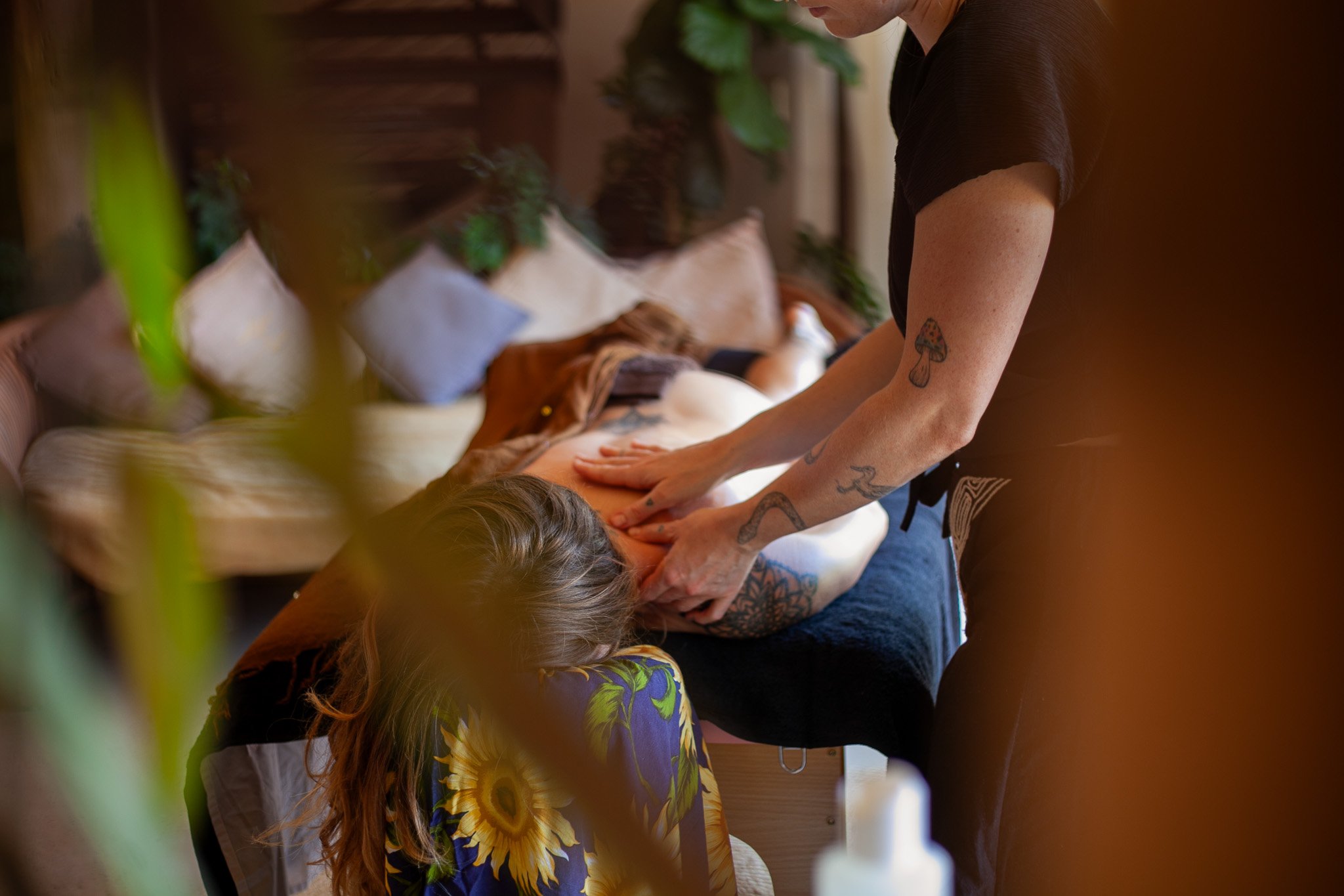 Person giving a massage to another person lying face down on a massage table, surrounded by a cozy, warmly lit room with pillows and plants.