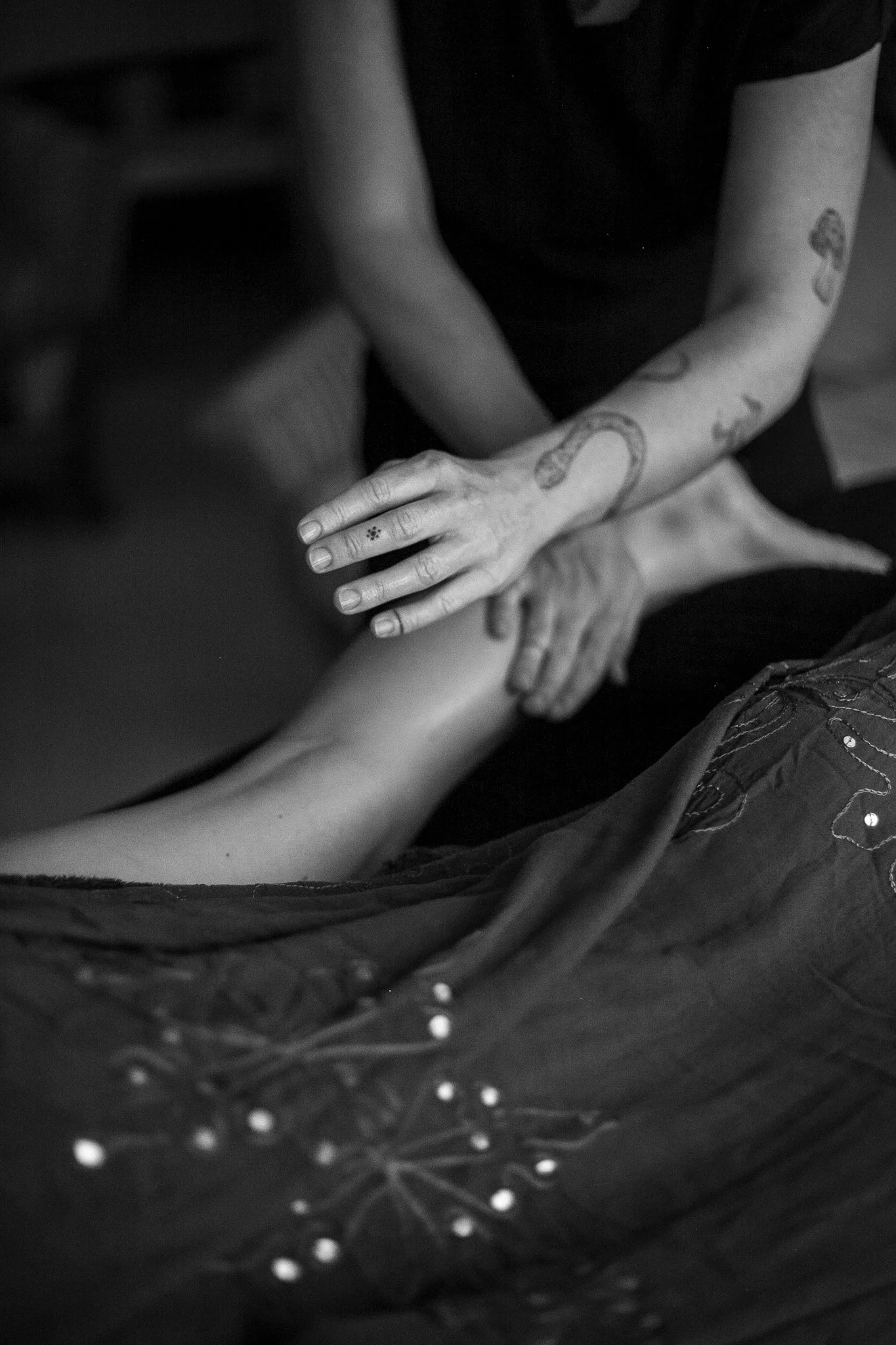 A person with tattoos on their arms gently touching another person's leg, in black and white.
