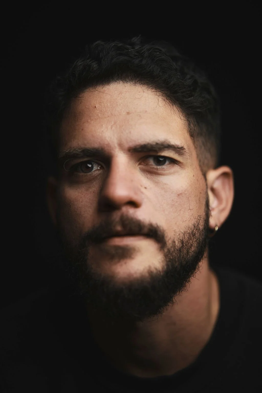 Close-up portrait of a man with dark hair and a beard, looking directly at the camera against a black background.