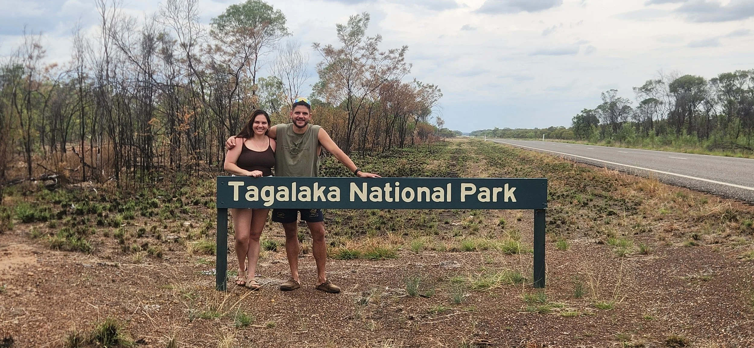 A couple standing behind a sign that reads "Tagalaka National Park" on a dirt path next to a road, with a background of sparse trees and overcast sky.