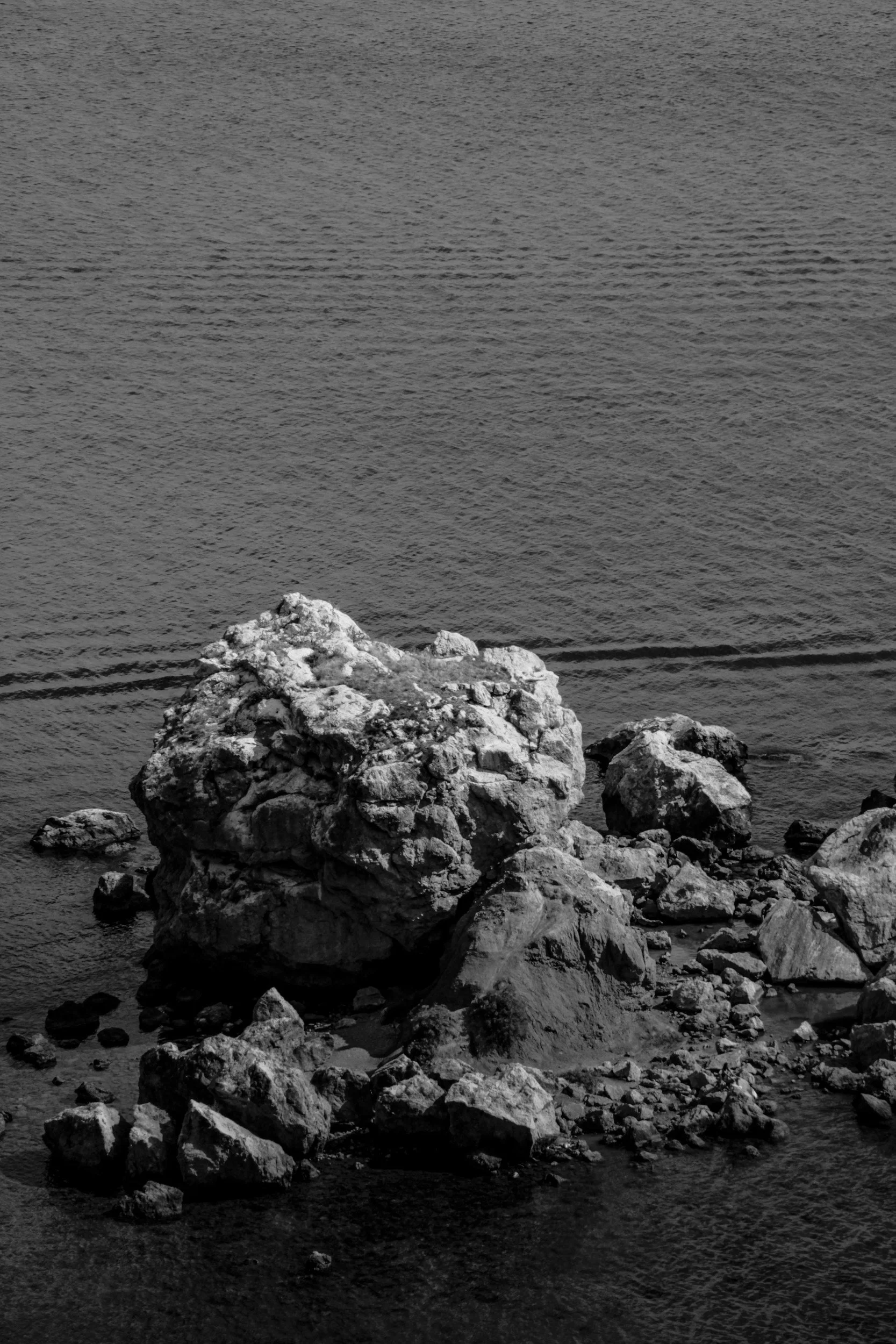 A black and white photograph of a large rocky formation in water, with smaller rocks surrounding it.