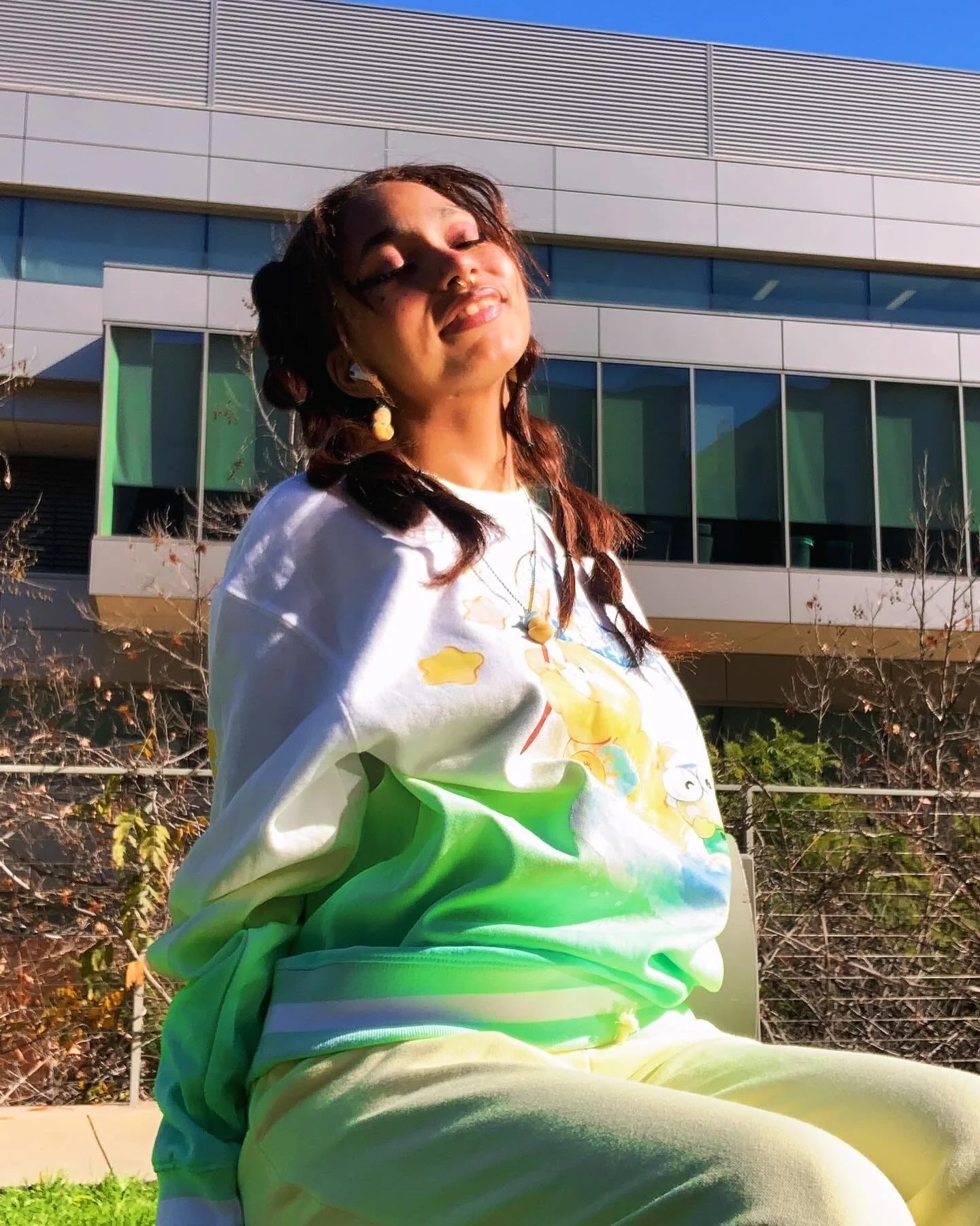 A woman with red hair and light brown skin, smiling with eyes closed, sitting outdoors in sunlight, in front of a modern building with large windows and a metal facade, wearing a colorful sweatshirt with cartoon characters and yellow pants.