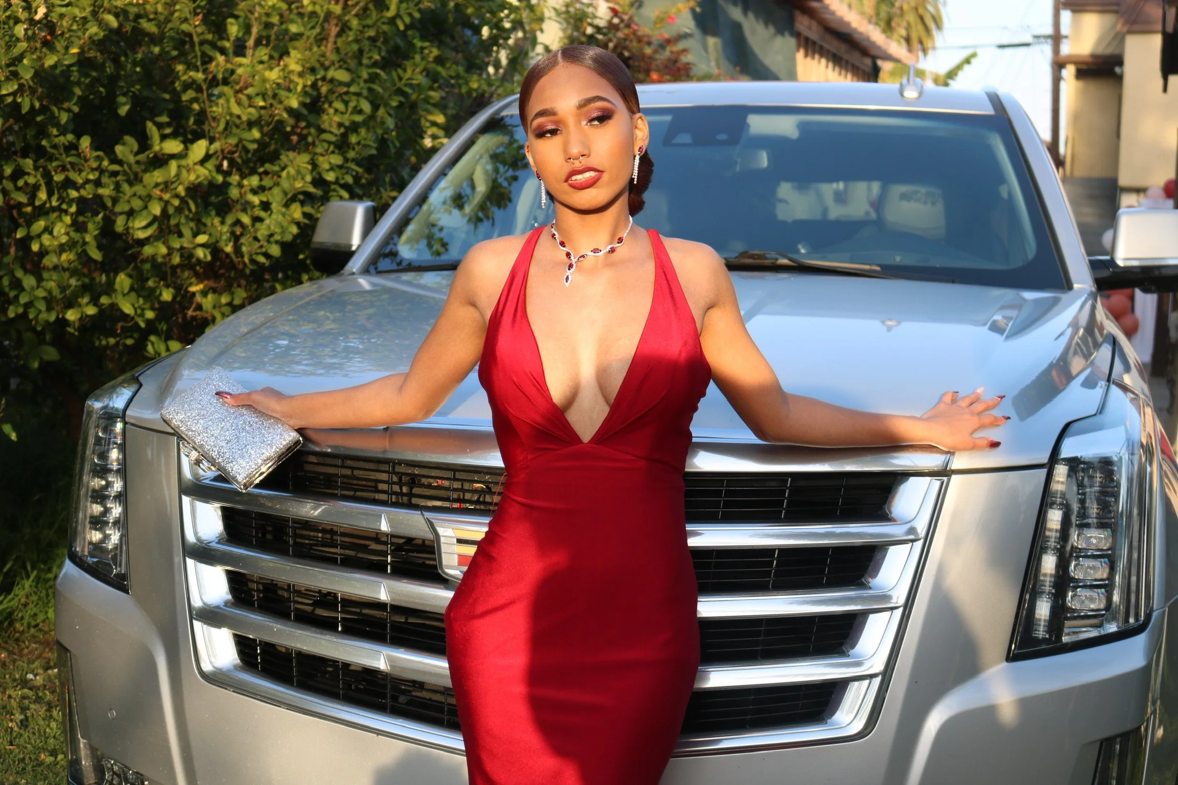 A woman in a red dress stands in front of a silver vehicle, holding a silver clutch bag, with greenery and buildings in the background.