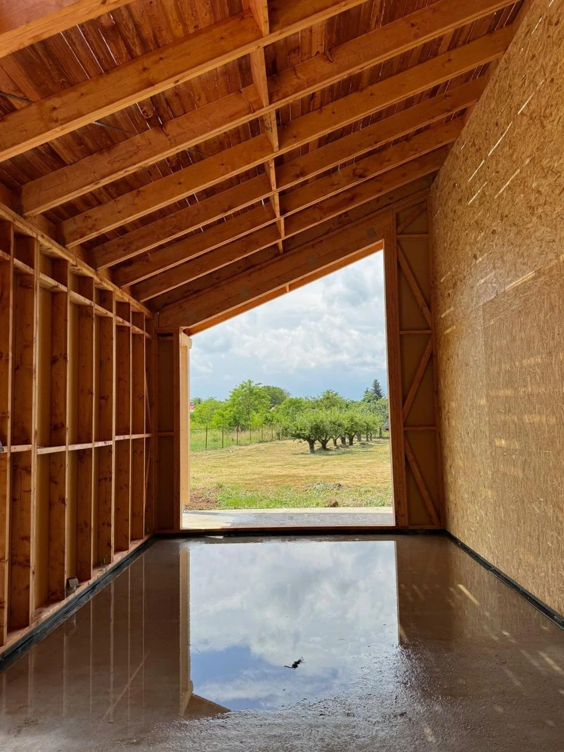 Interior of a wooden structure under construction with an open front view showing trees and a cloudy sky outside, a polished concrete floor reflecting the sky.