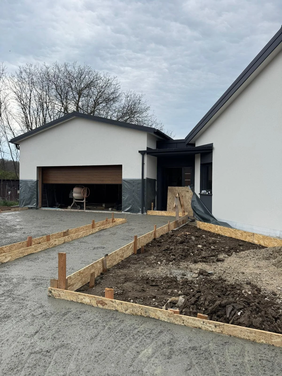 Exterior of a house under construction, with a driveway being prepared, wooden forms for concrete, and a garage door.