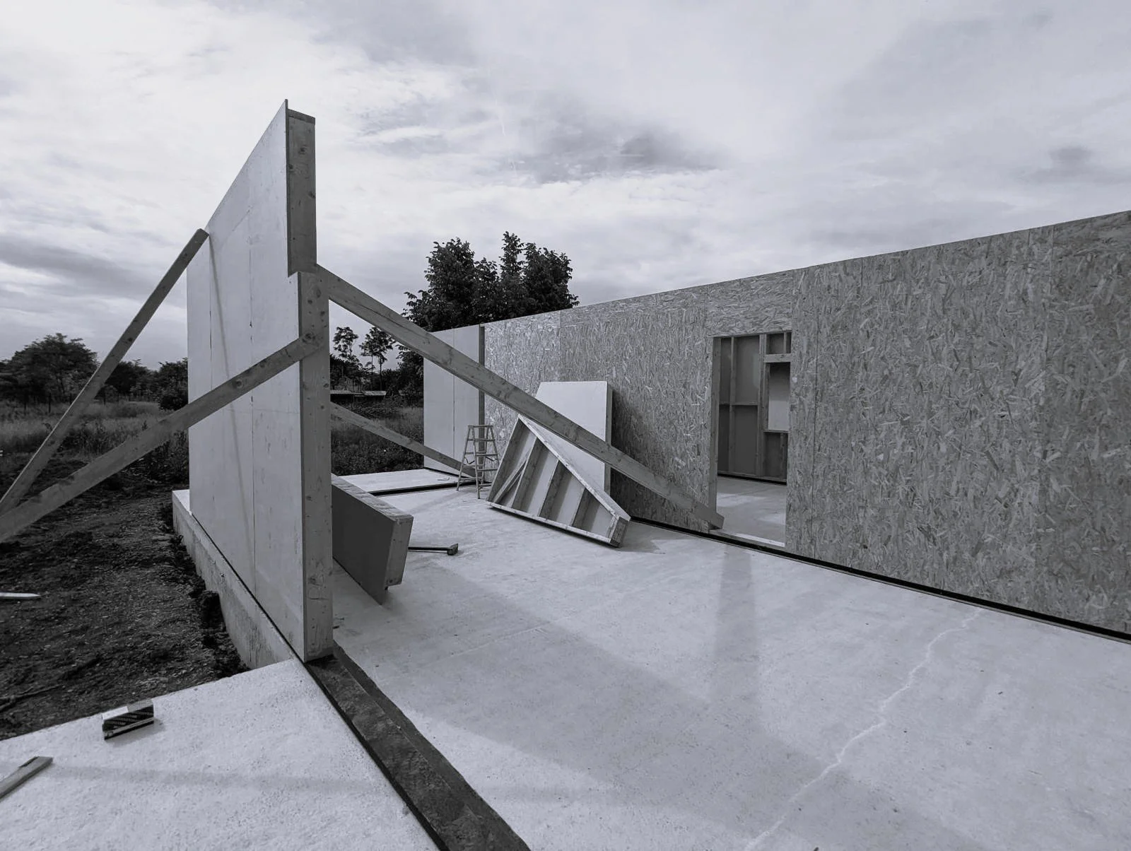 Construction site with partially built walls of a building, including plywood and framed sections, on a concrete floor with tools and a ladder, under a cloudy sky.