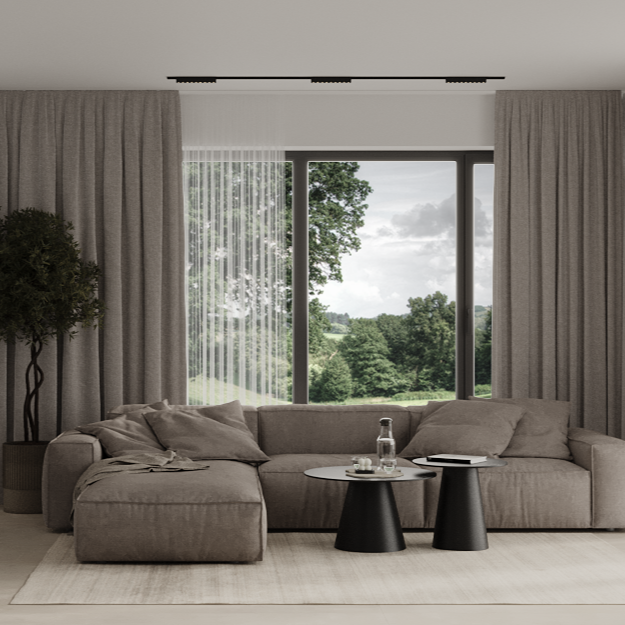 Modern living room with beige sofa, two black coffee tables, a glass of water, a remote, and three pillows, overlooking a lush green landscape through large windows.