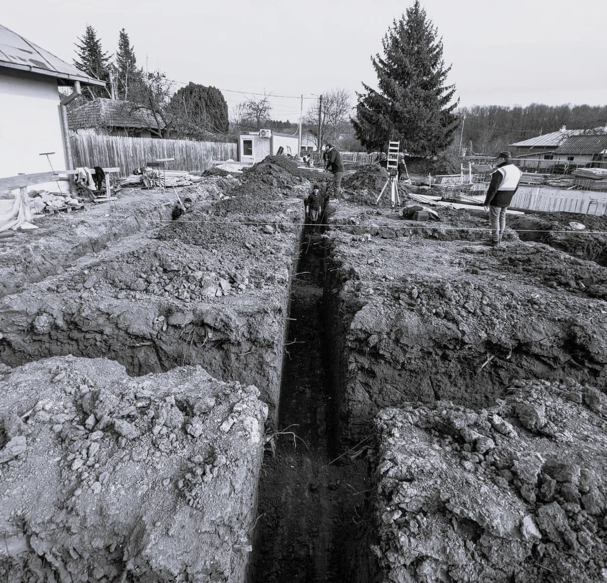 Construction workers digging a trench at a construction site in a residential area with houses and trees in the background.