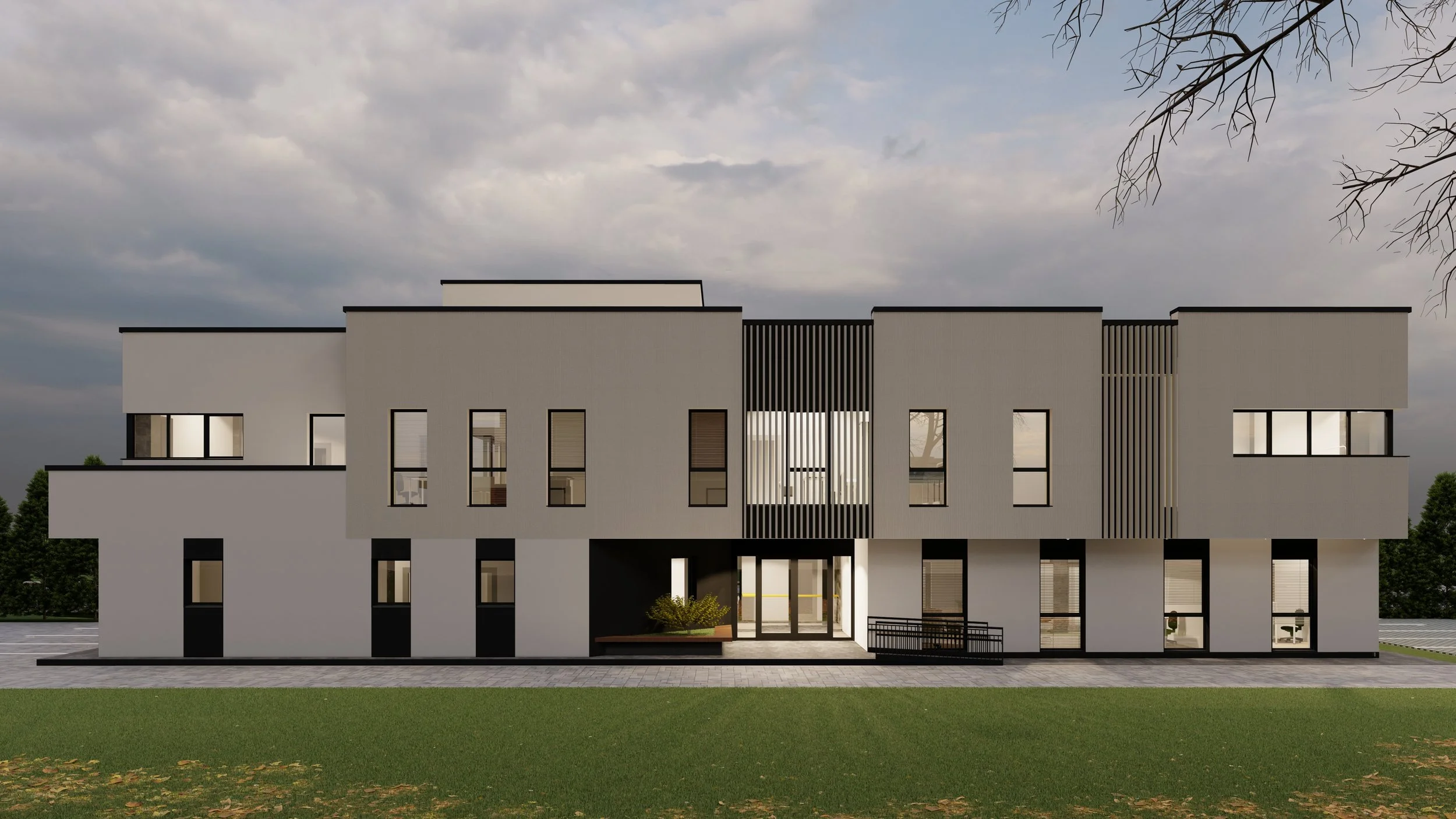 Modern multi-story building with white walls and black accents, featuring large windows and a central entrance with a small landscaped area, surrounded by a green lawn and trees in the background under a partly cloudy sky.