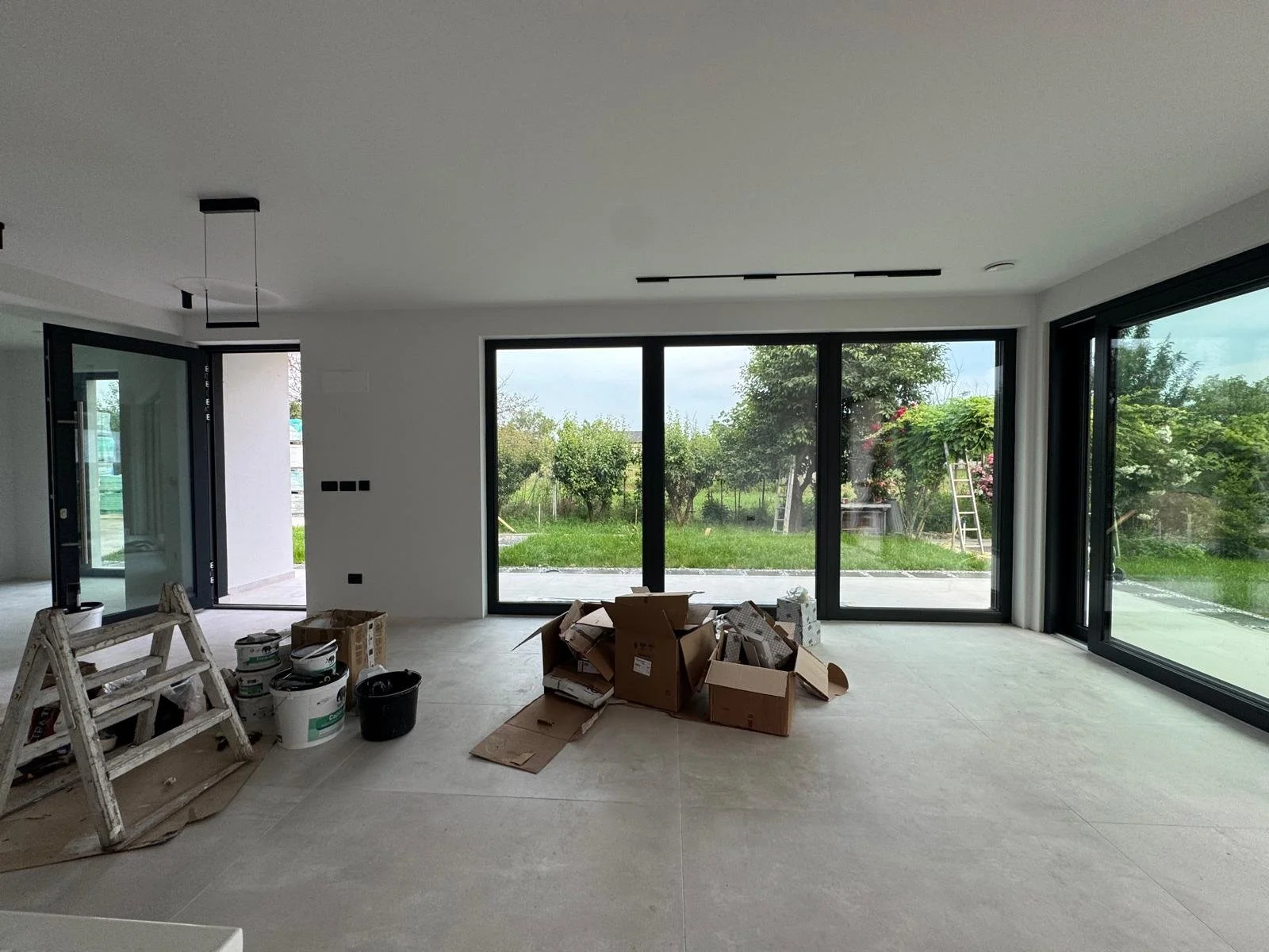 Interior of a modern room under construction with large sliding glass doors and view of a garden outside. Construction materials and tools are scattered on the floor.