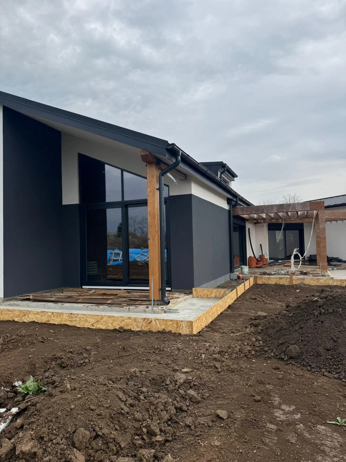 Under construction modern house with dark gray walls, large glass doors, and wooden framework on the patio.