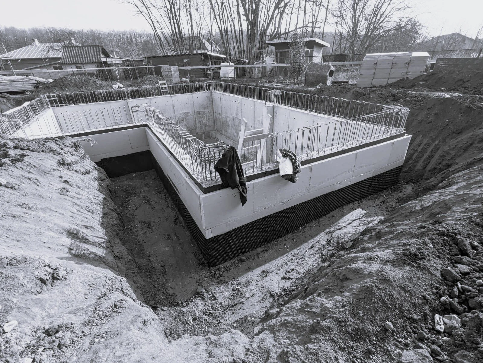 Construction site showing the foundation of a building with concrete walls and rebar, surrounded by excavated dirt and debris.