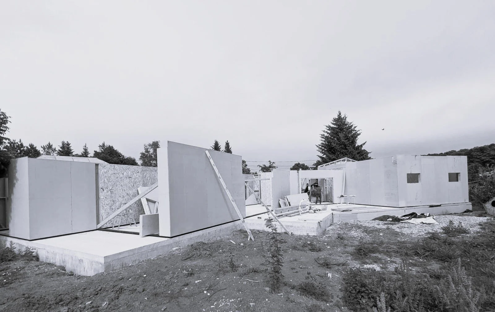 Construction site with partially built white walls and framing, surrounded by dirt and construction materials, with trees and cloudy sky in the background.