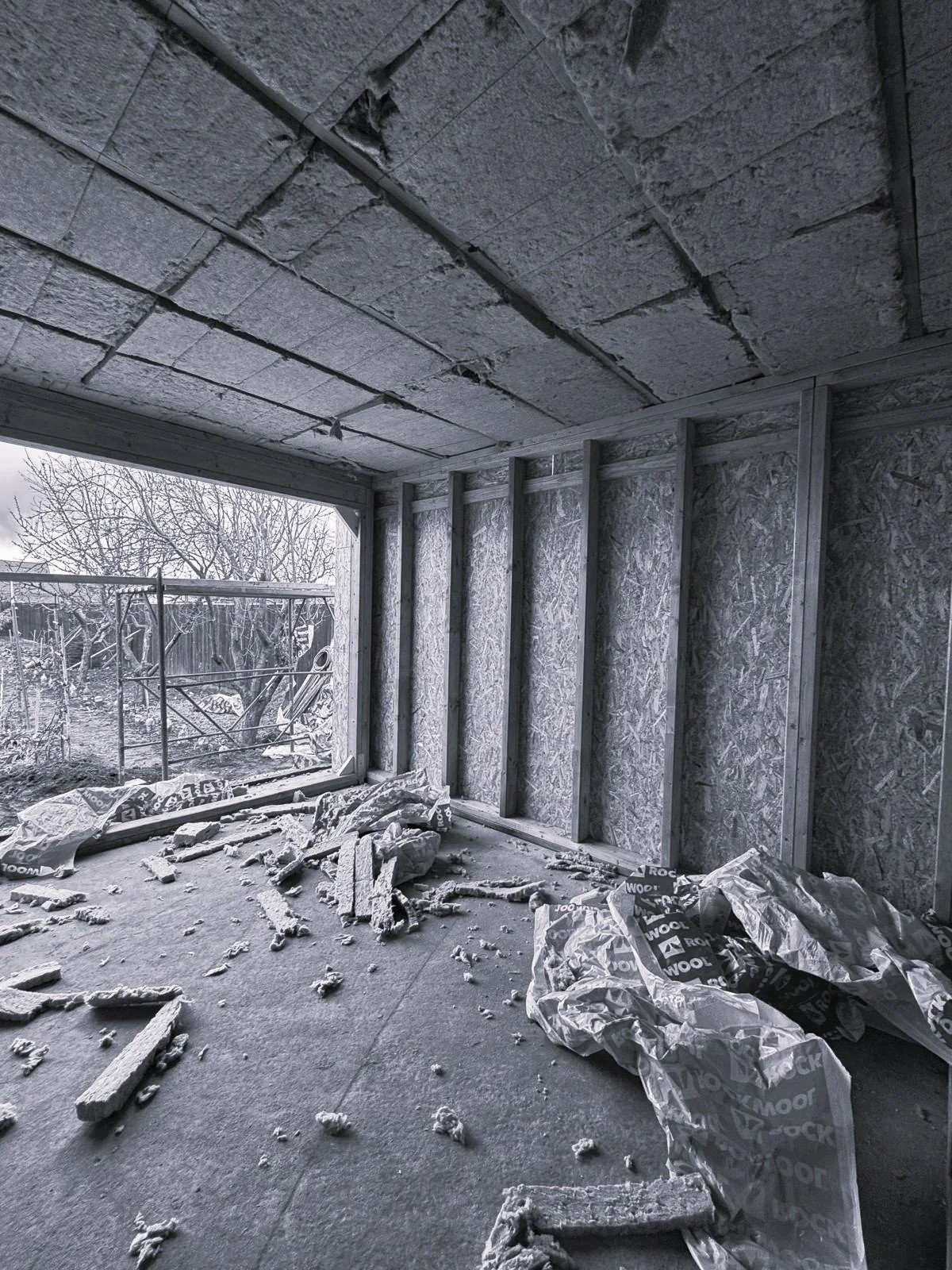 Interior of a building under construction with exposed insulation, wooden framing, and debris on the floor.