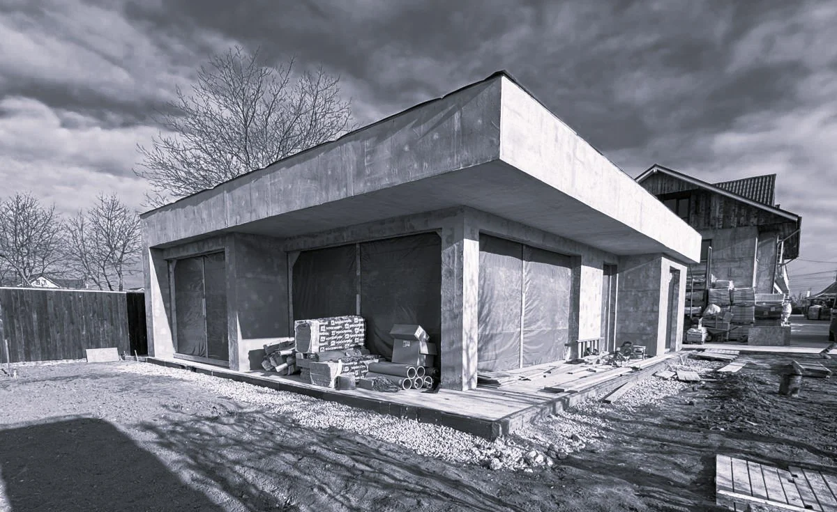 Under-construction modern house with brick and concrete exterior, construction materials like bricks, pipes, and wood scattered around, cloudy sky, and other buildings in the background.