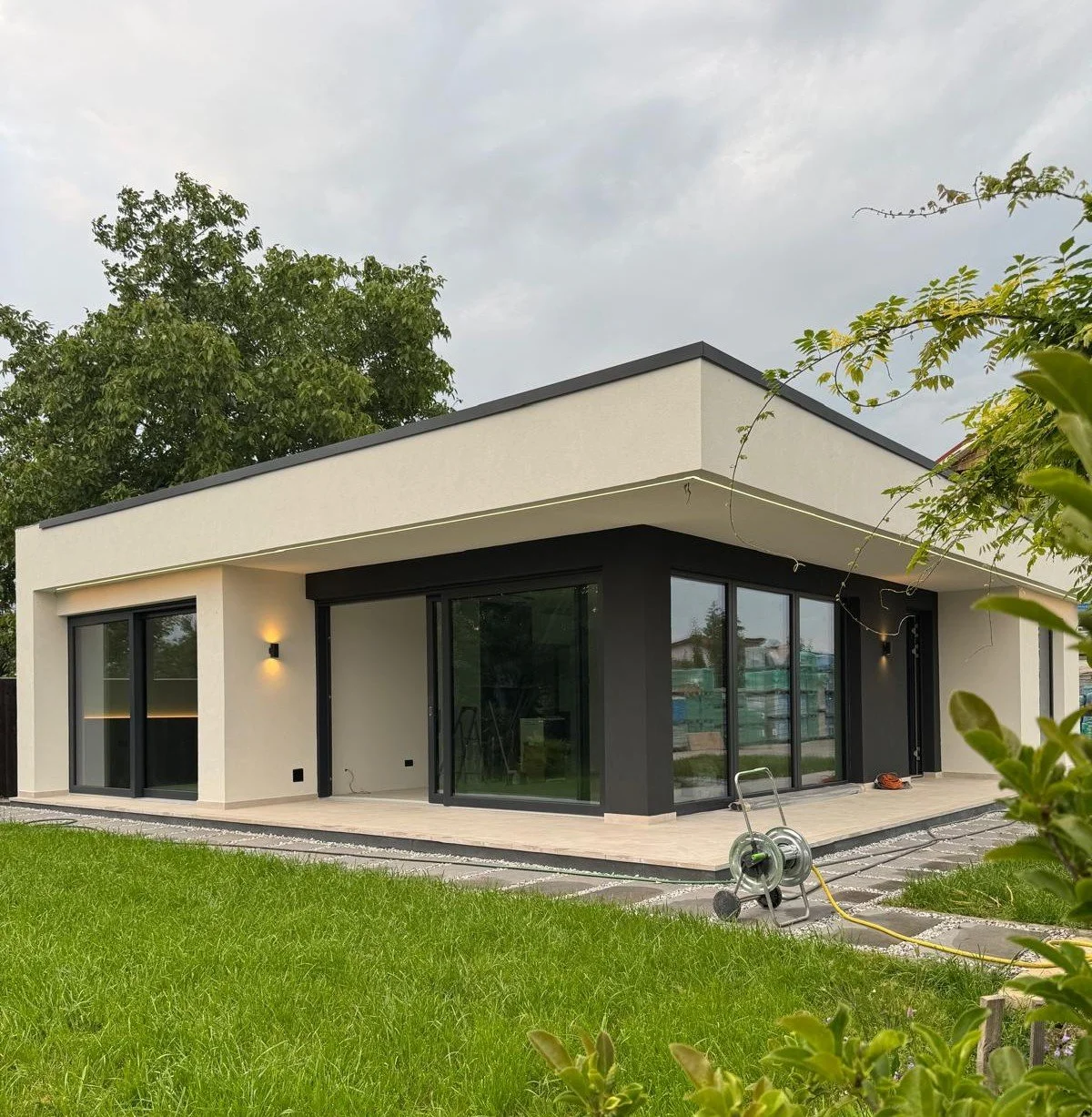 Modern white house with large black-framed glass sliding doors and windows, surrounded by green grass and trees, under an overcast sky.