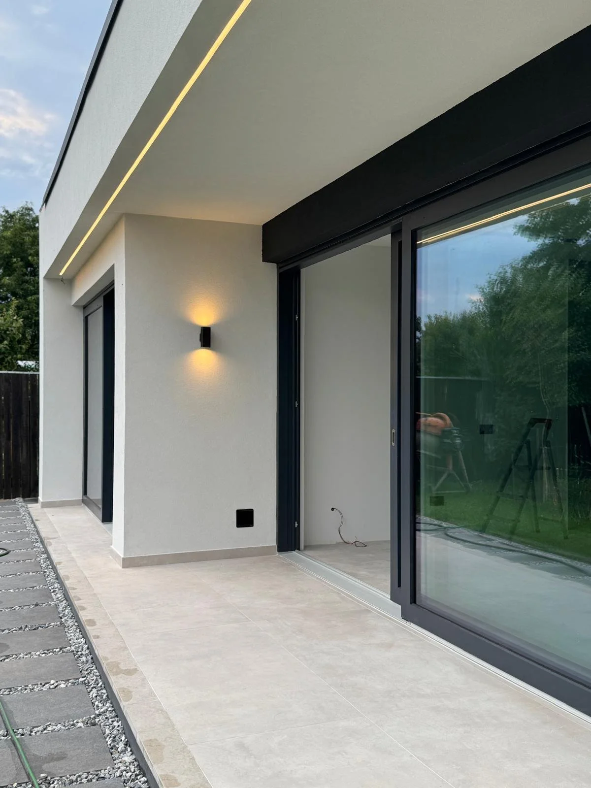 Modern house exterior with white walls, sliding glass doors, outdoor tiled patio, black wall-mounted light, and a fence with trees in the background.