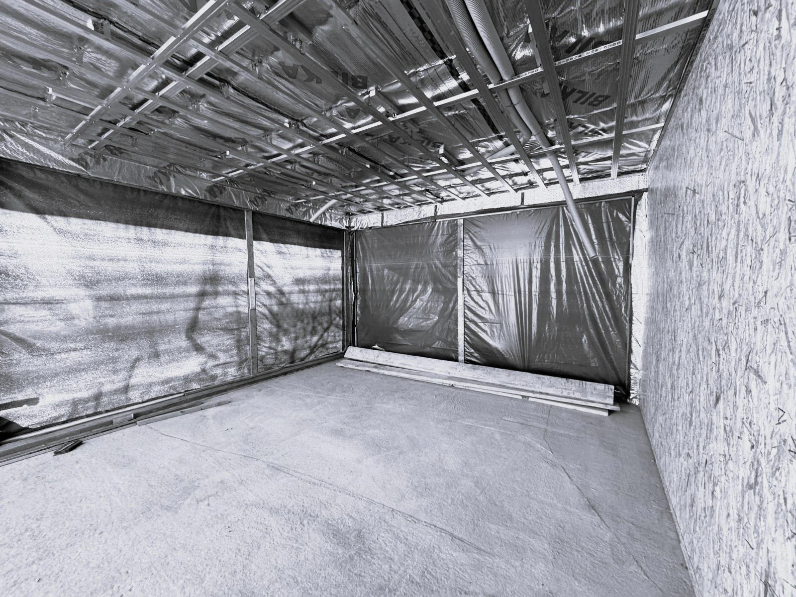 Construction site with framed walls, exposed ceiling with pipes and wires, and black plastic sheeting covering parts of the walls.