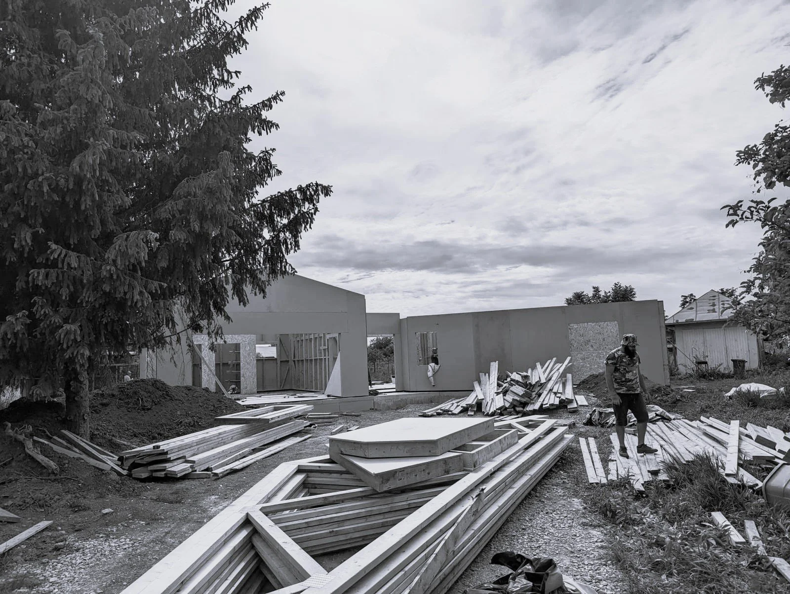 Construction site with building walls, scattered wood planks, and a worker wearing a hat and camouflage shirt, under a cloudy sky.