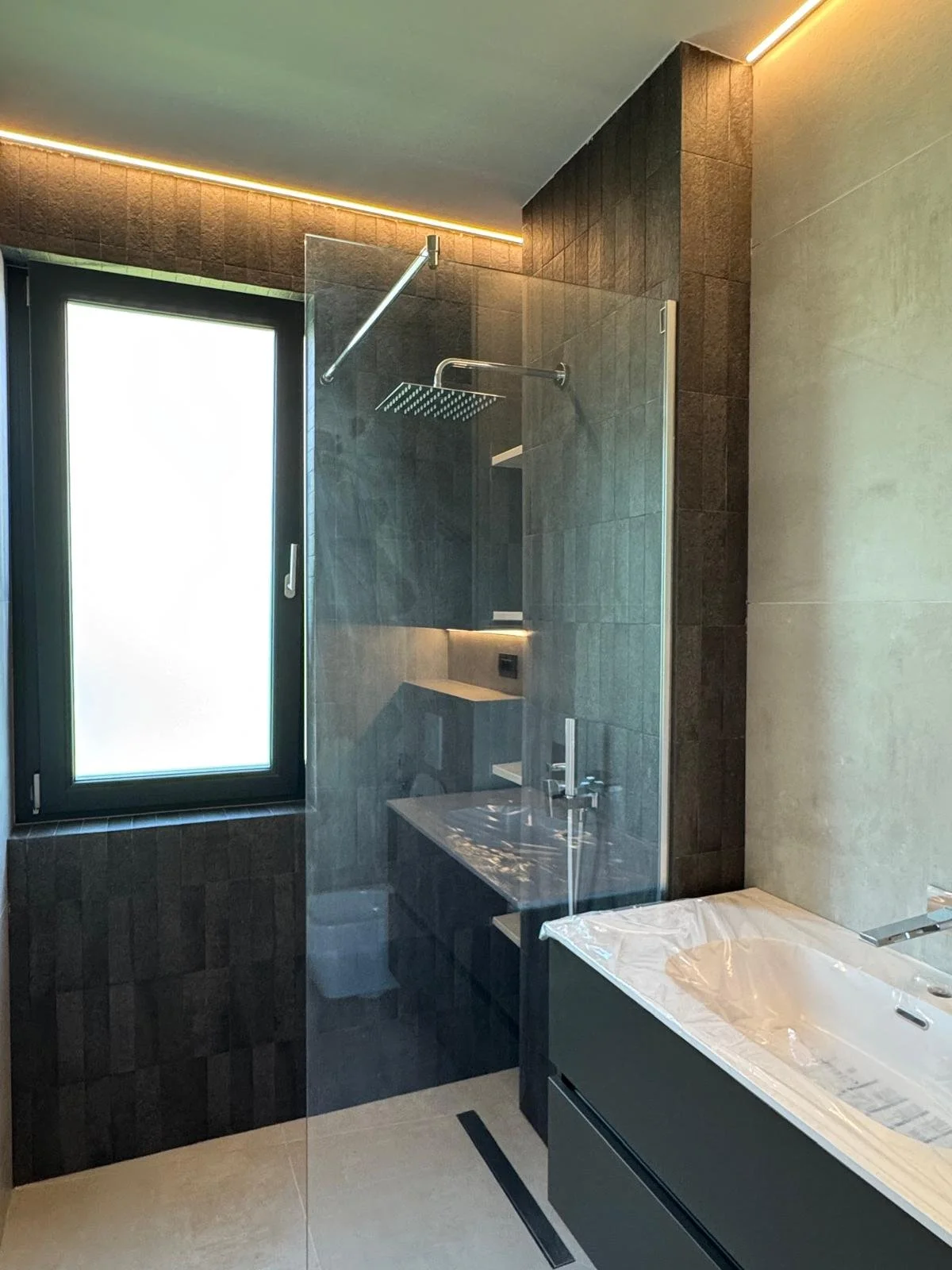 Modern bathroom with dark tiled shower area, glass partition, white sink, and frosted window, illuminated by warm strip lighting.
