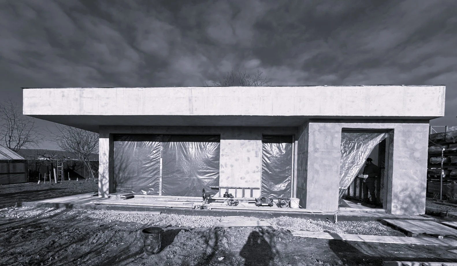 Construction site with a partially built modern concrete house, worker on site, tools and materials scattered on ground, cloudy sky overhead.