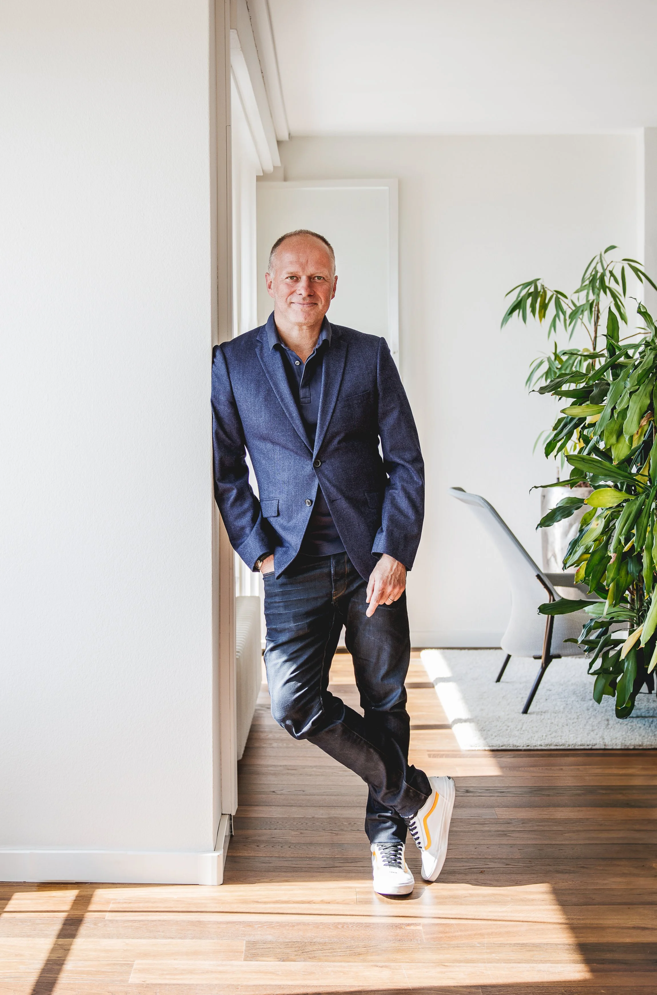 Un homme debout dans une pièce lumineuse, portant une veste bleue, un pantalon en jean, et des baskets blanches, posant près d'un mur blanc avec une plante verte à côté.