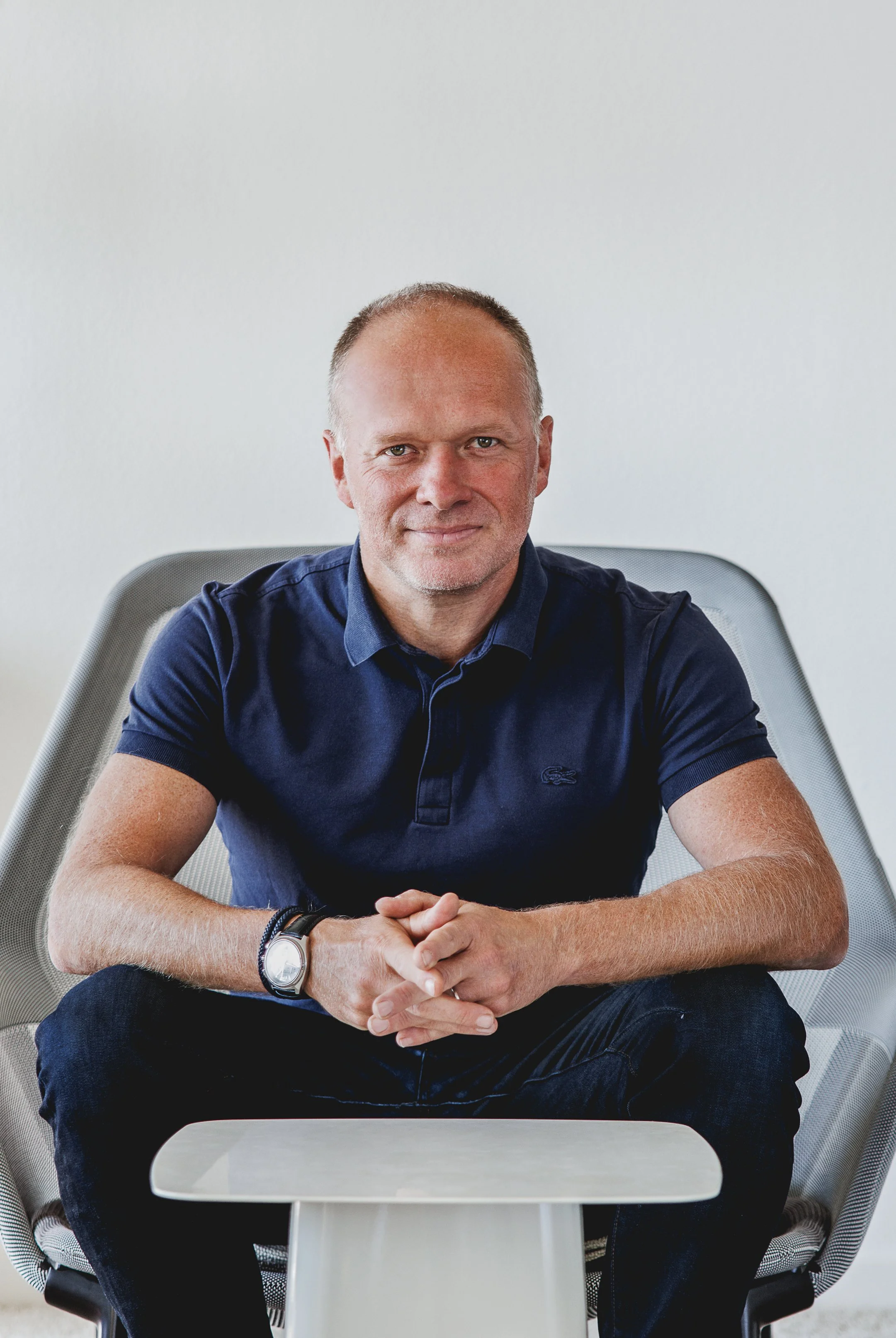 Un homme assis à une table, portant une chemise bleue, souriant légèrement, dans un environnement intérieur simple avec un fond blanc et une chaise moderne.