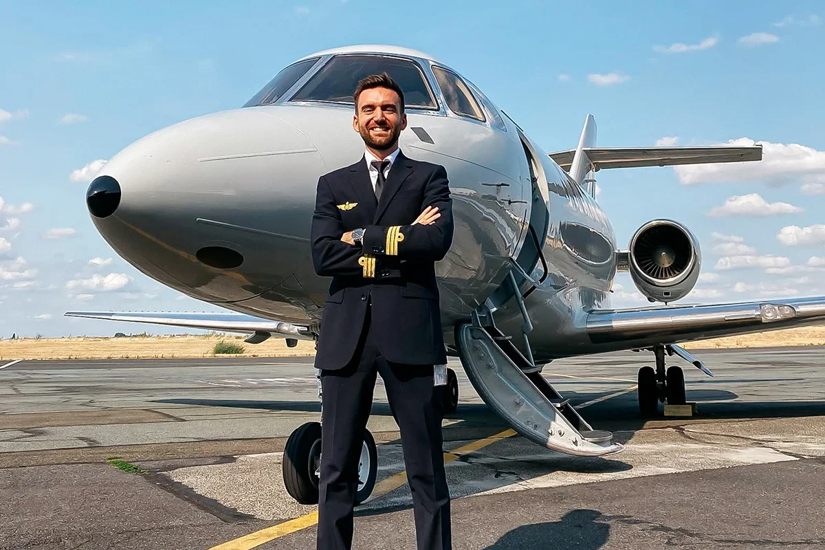 Un pilote en tenue formelle debout devant un jet privé sur un tarmac avec un ciel bleu clair et quelques nuages.