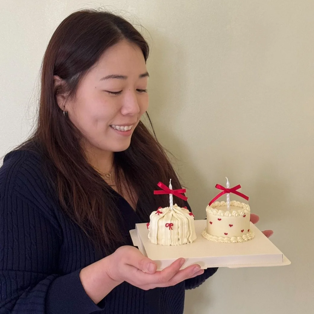A woman with long dark hair smiling while holding a tray with two small decorated cakes, each with a candle and red ribbon bows, against a plain beige wall.