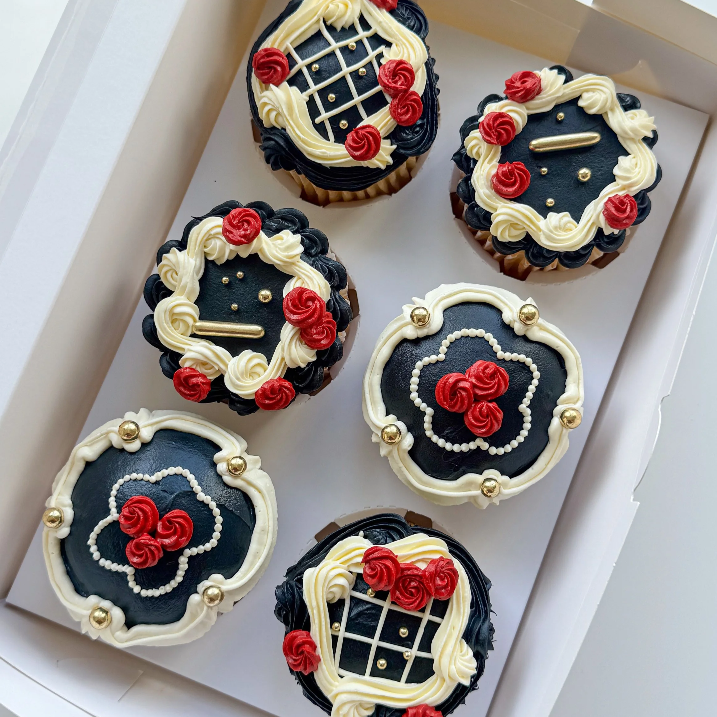 Six decorated cupcakes with black, white, and red icing, adorned with gold and white edible beads, in a white box.