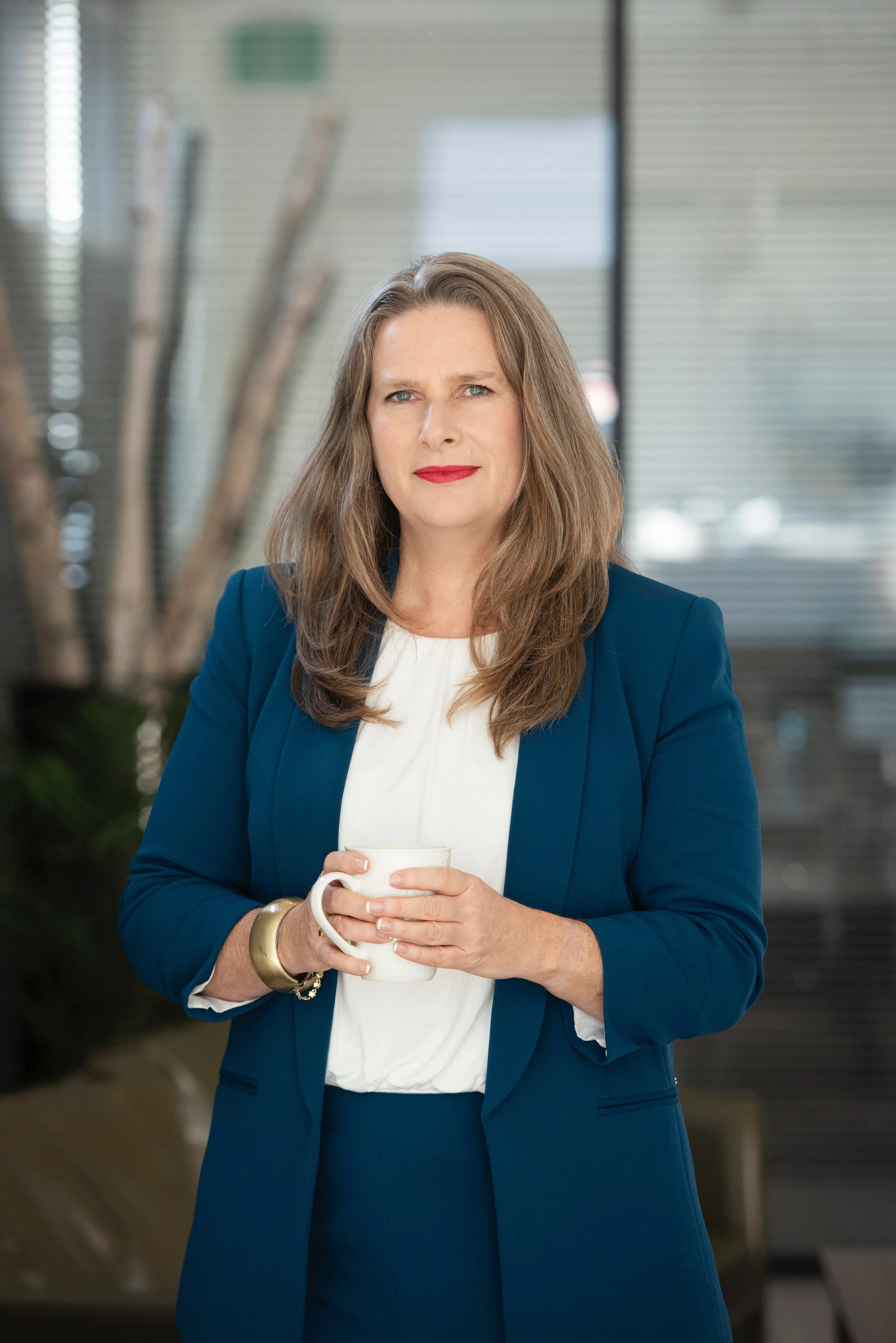 A woman with long brown hair, wearing a blue blazer and white blouse, holding a white coffee mug, standing in an office setting.