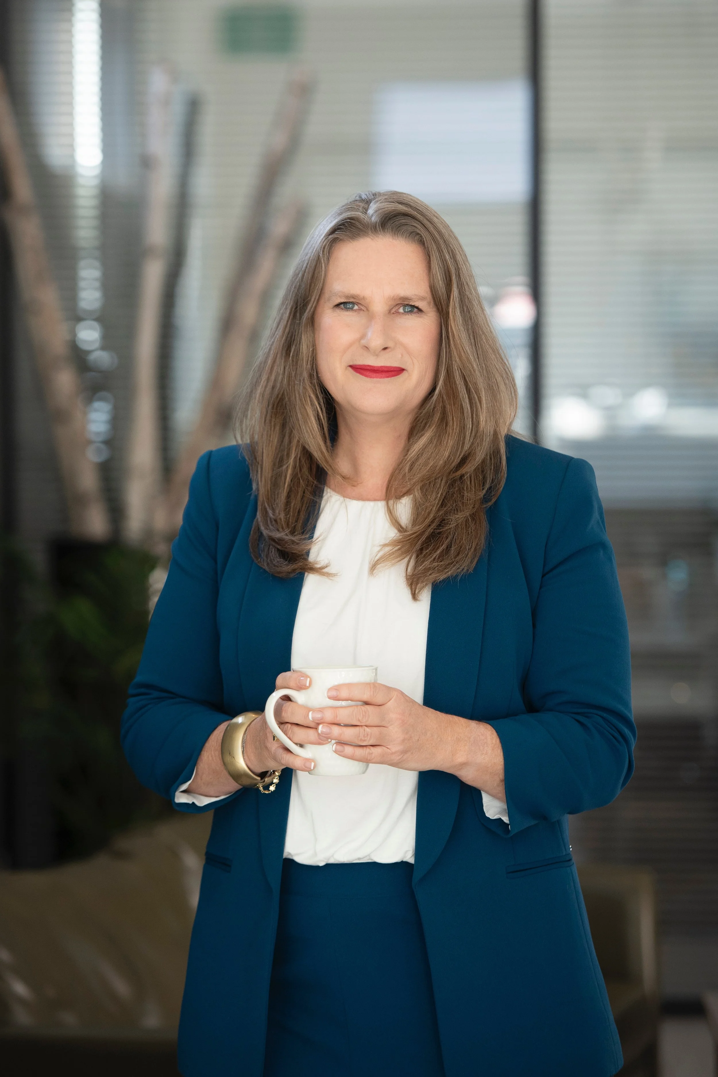 A professional woman with long, wavy brown hair wearing a navy blazer and white blouse, holding a white coffee mug, standing indoors with a blurred modern office background.