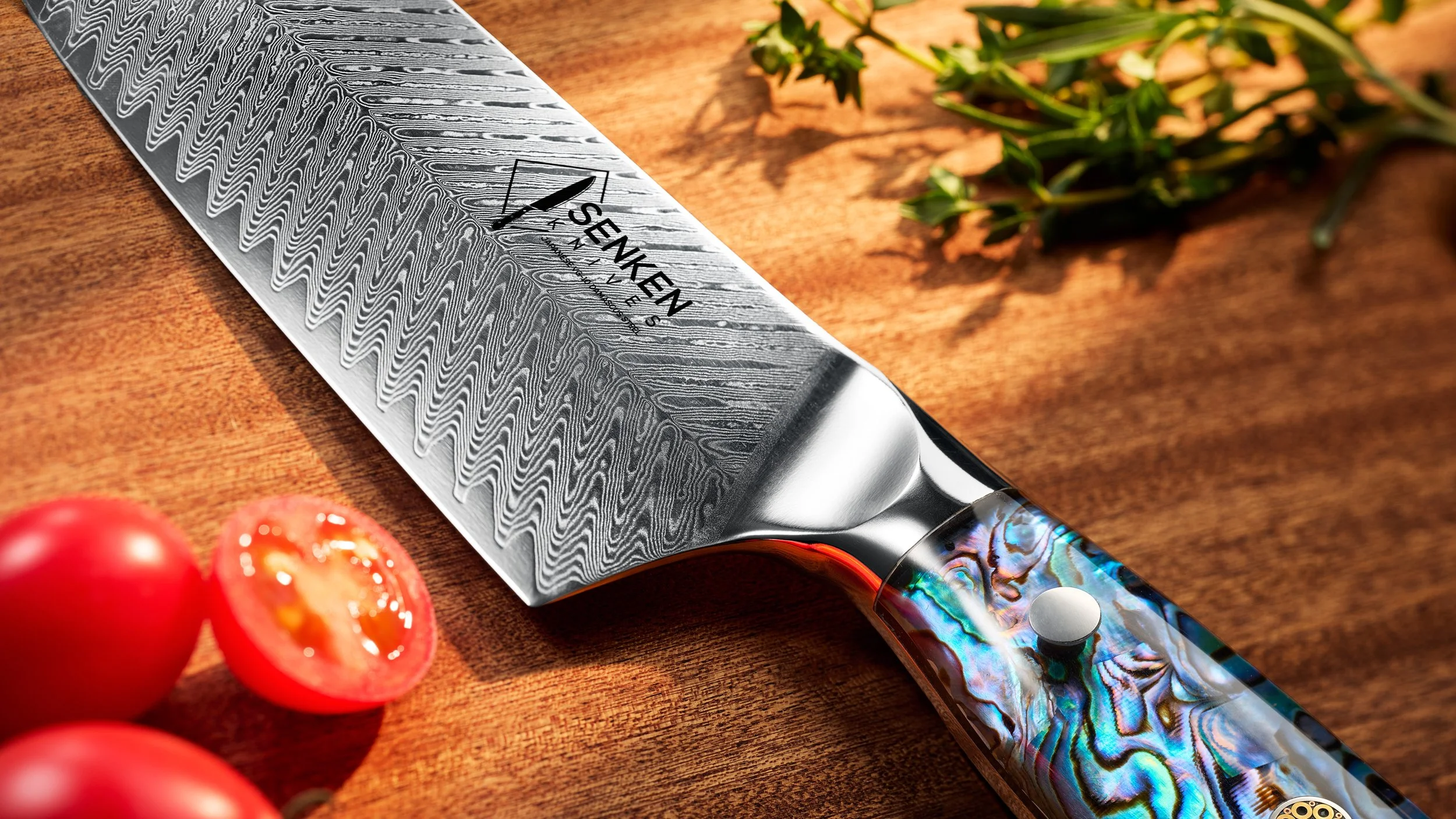 Close-up of a Damascus steel kitchen knife with a handle made of colorful abalone shell, resting on a wooden surface. Fresh tomatoes and herbs are also visible nearby.