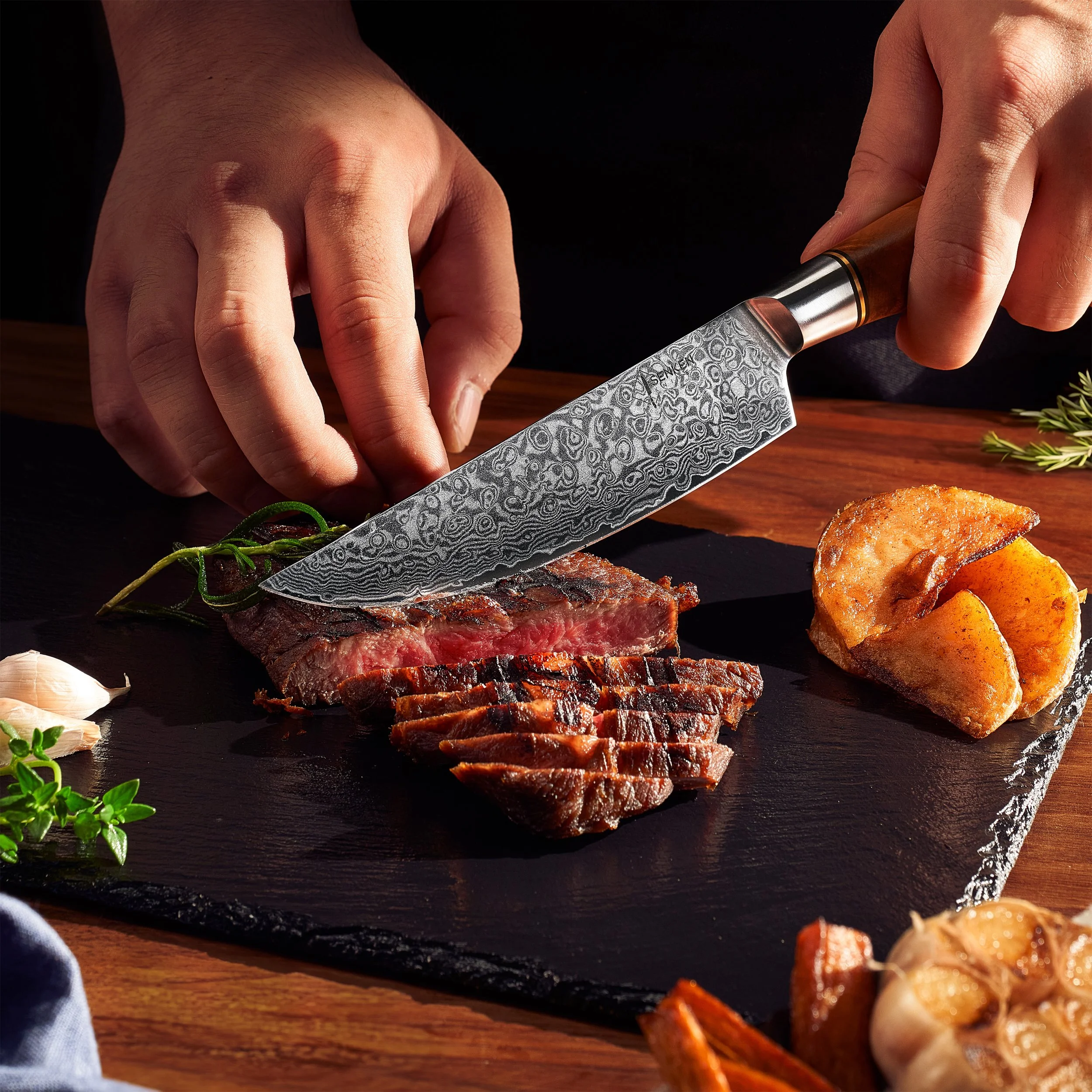A person slicing a cooked steak on a black slate cutting board, with roasted potatoes, garlic, and herbs nearby.