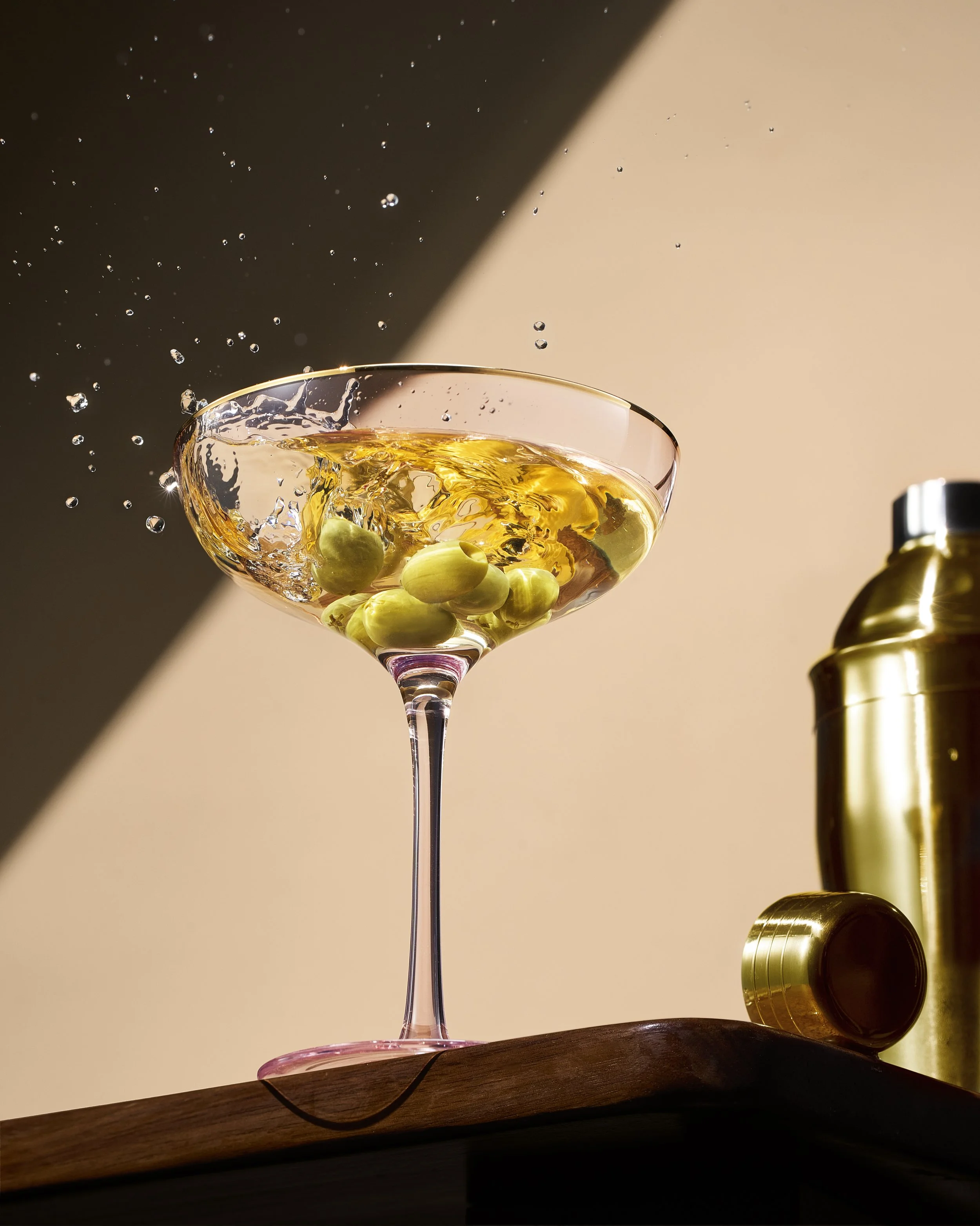 A cocktail glass with green olives being poured with a golden drink, with bubbles splashing out and a cocktail shaker on the right.