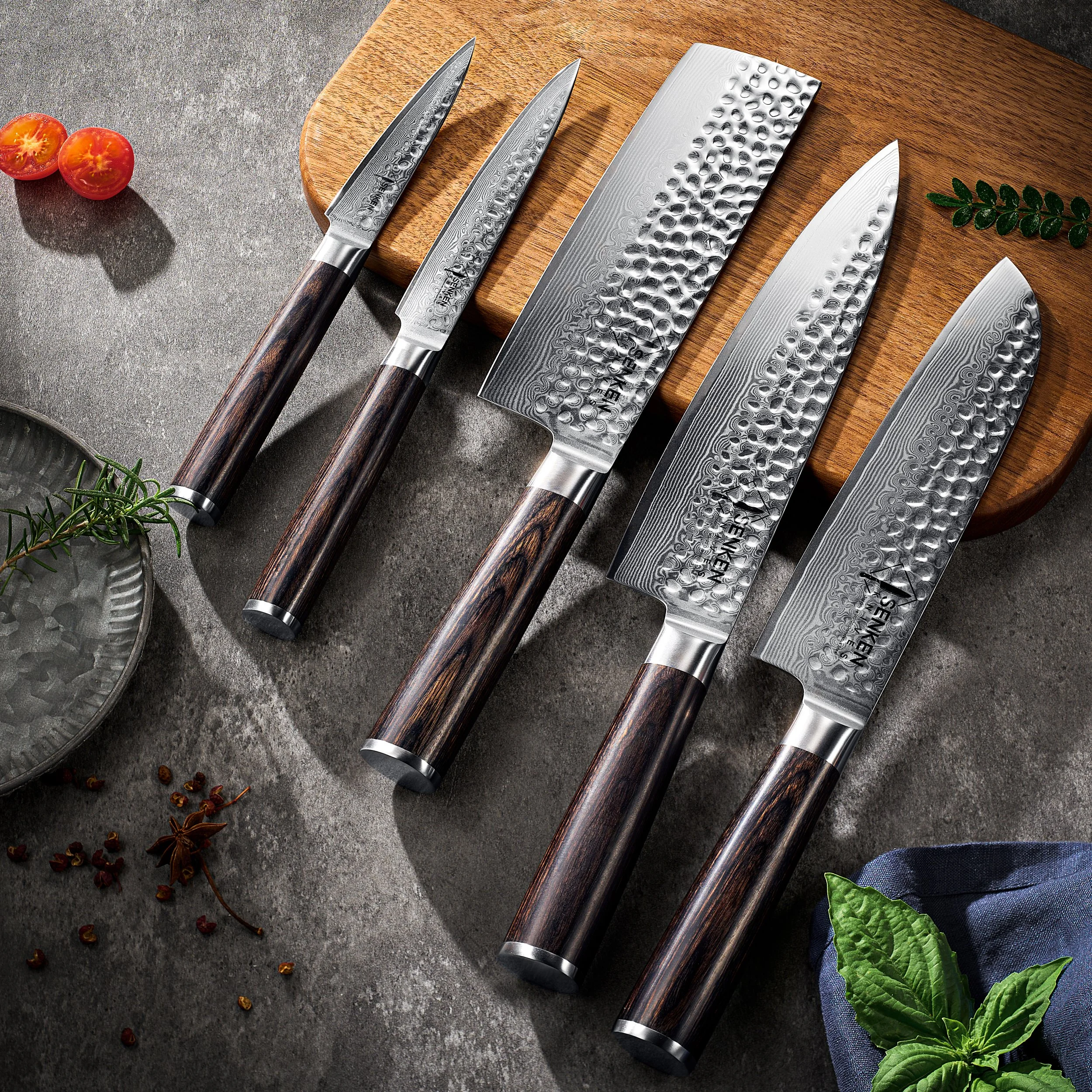 Set of five Japanese-style kitchen knives with hammered metal blades and dark wooden handles, arranged on a gray surface with a wooden cutting board, cherry tomatoes, herbs, and spices nearby.